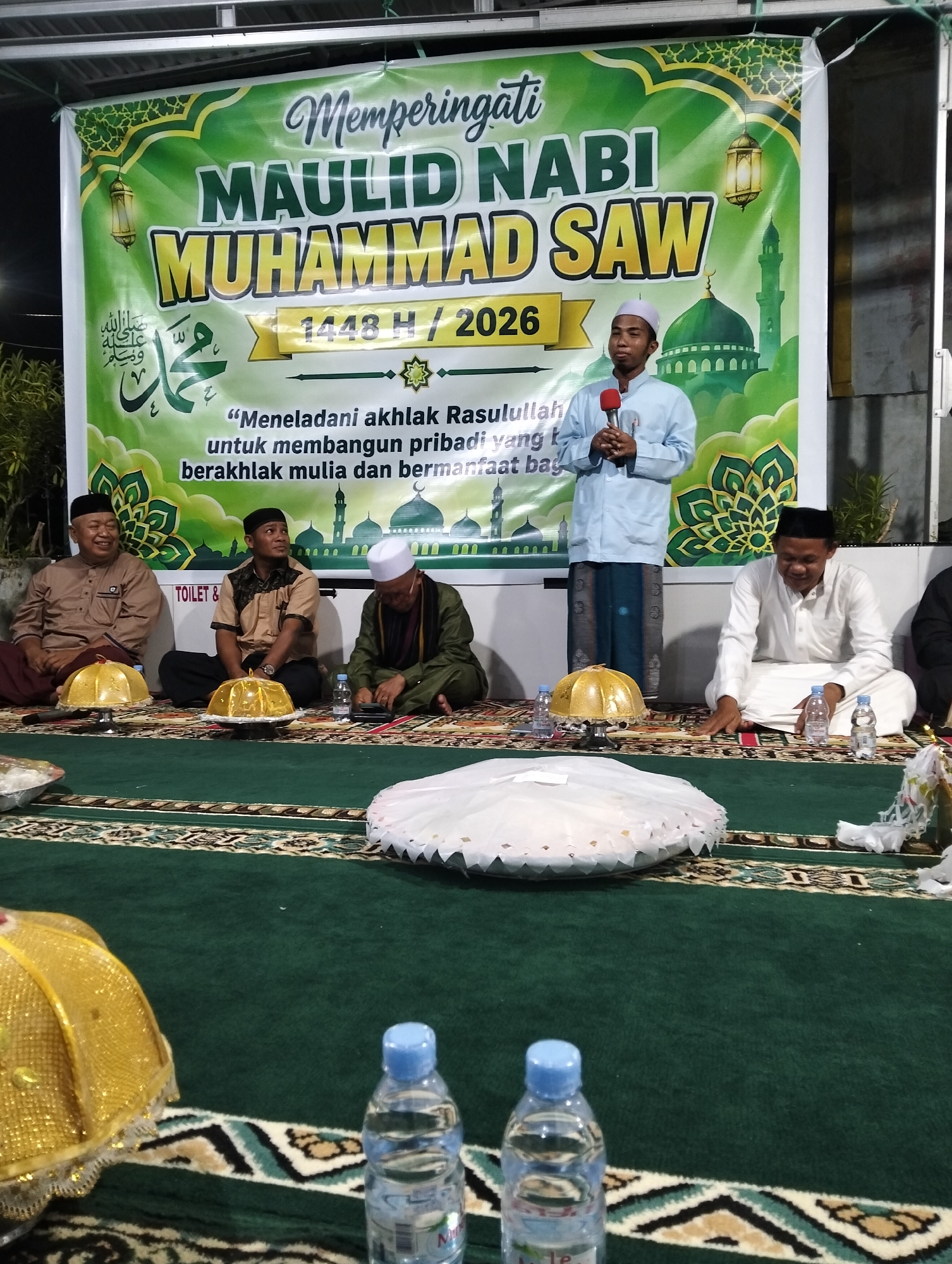 The image depicts an indoor event, likely a religious gathering, as indicated by the banner in the background celebrating "MAULID NABI MUHAMMAD SAW," which translates to the "Birthday of Prophet Muhammad SAW." The banner features Islamic calligraphy and imagery of mosques and lanterns, common in Muslim celebrations.
In the foreground, a green carpet covers the floor, adorned with a patterned border. Several men are seated on the carpet, dressed in traditional Islamic attire. One man in the center is speaking into a microphone, suggesting he is a speaker or imam. He is wearing a light blue shirt and a white cap.
To his left, three men are seated. The man on the far left is wearing a brown shirt and a maroon sarong, with a wide smile. Next to him is a man in a brown and beige patterned shirt and black pants. The third man is wearing a white cap, a dark green robe, and a patterned scarf. He is looking down at a book or device.
To the right of the speaker, a man in a white shirt and white pants is seated, looking to his left with a smile. Further to the right, partially visible, is another man in dark clothing.
Several gold-domed decorative objects are placed on the carpet in front of the seated men. Bottles of water are also visible on the carpet. In the background, a white banner with green decorative elements is hung. The overall scene suggests a formal gathering with a spiritual or religious theme, likely held in a community hall or similar venue. There is no explicit indication of the specific location within Petapa, Indonesia, beyond it being a local event.