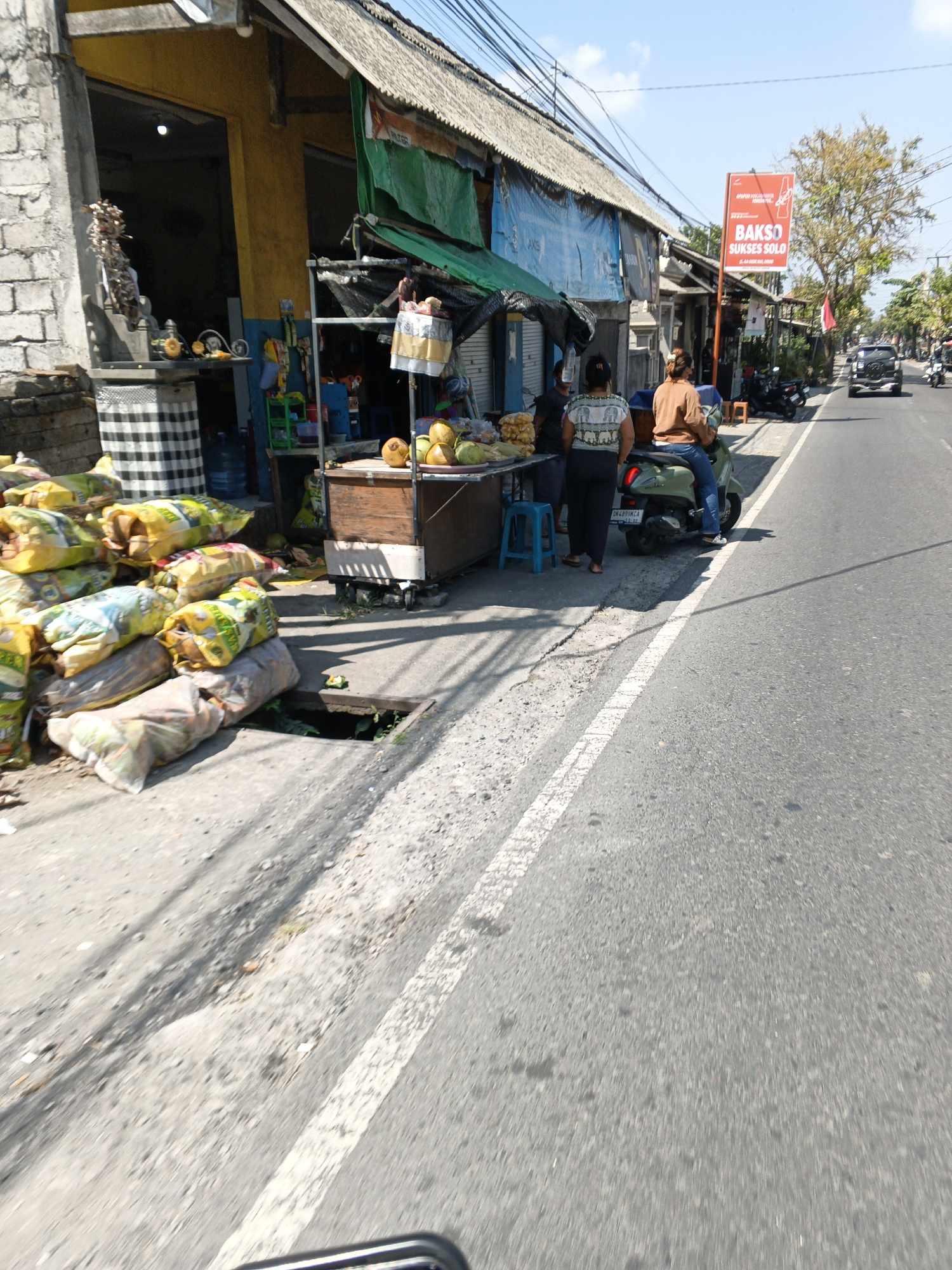 This is a street-level, medium-distance, eye-level shot of a roadside stall and street scene in Ubud District, Indonesia. The stall appears to be selling food items, with large bags of produce or goods stacked on the left and a wooden cart displaying coconuts and other items. Two women are standing at the stall, one in a white patterned top and dark pants, and another in a brown long-sleeved shirt and blue jeans, seated on a green scooter. A man in a brown shirt and blue jeans is also seated on the scooter behind the woman. The street is paved with asphalt and has a white line marking a lane. A red sign with the text "BAKSO SUKSES SOLO" is visible in the background, along with a Coca-Cola logo. The sky is clear and blue. Electrical wires are strung overhead. The overall impression is that of a busy, open-air market or food stand on a street in a tropical environment.