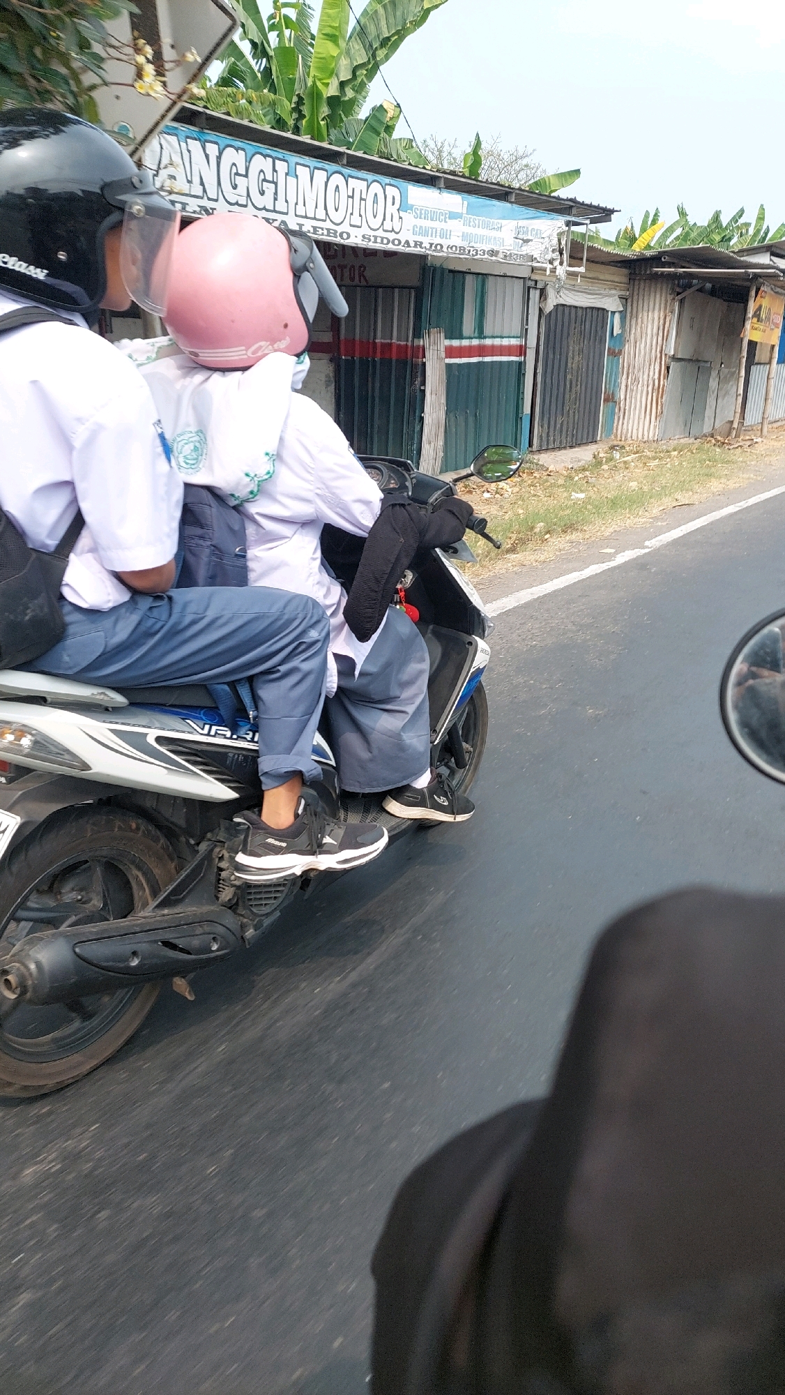 Two individuals, likely students given their white shirts and blue pants, are riding on a scooter. The person in the rear is wearing a pink helmet and appears to be a female. The person in the front is wearing a black helmet. The scooter is traveling on a road in front of a row of shops. One shop's signage reads "ANGGI MOTOR" and includes text that appears to be in Indonesian, with words like "SERVICE", "RESTORASI", and "MODIFIKASI", suggesting it is a motorcycle repair and customization shop. The road appears to be paved, and the environment suggests a warm, possibly tropical climate with visible greenery. The presence of "SIDOARJO" in the shop's signage clearly places the location in the city of Sidoarjo, Indonesia. The perspective suggests the photo was taken from another vehicle traveling behind the scooter.