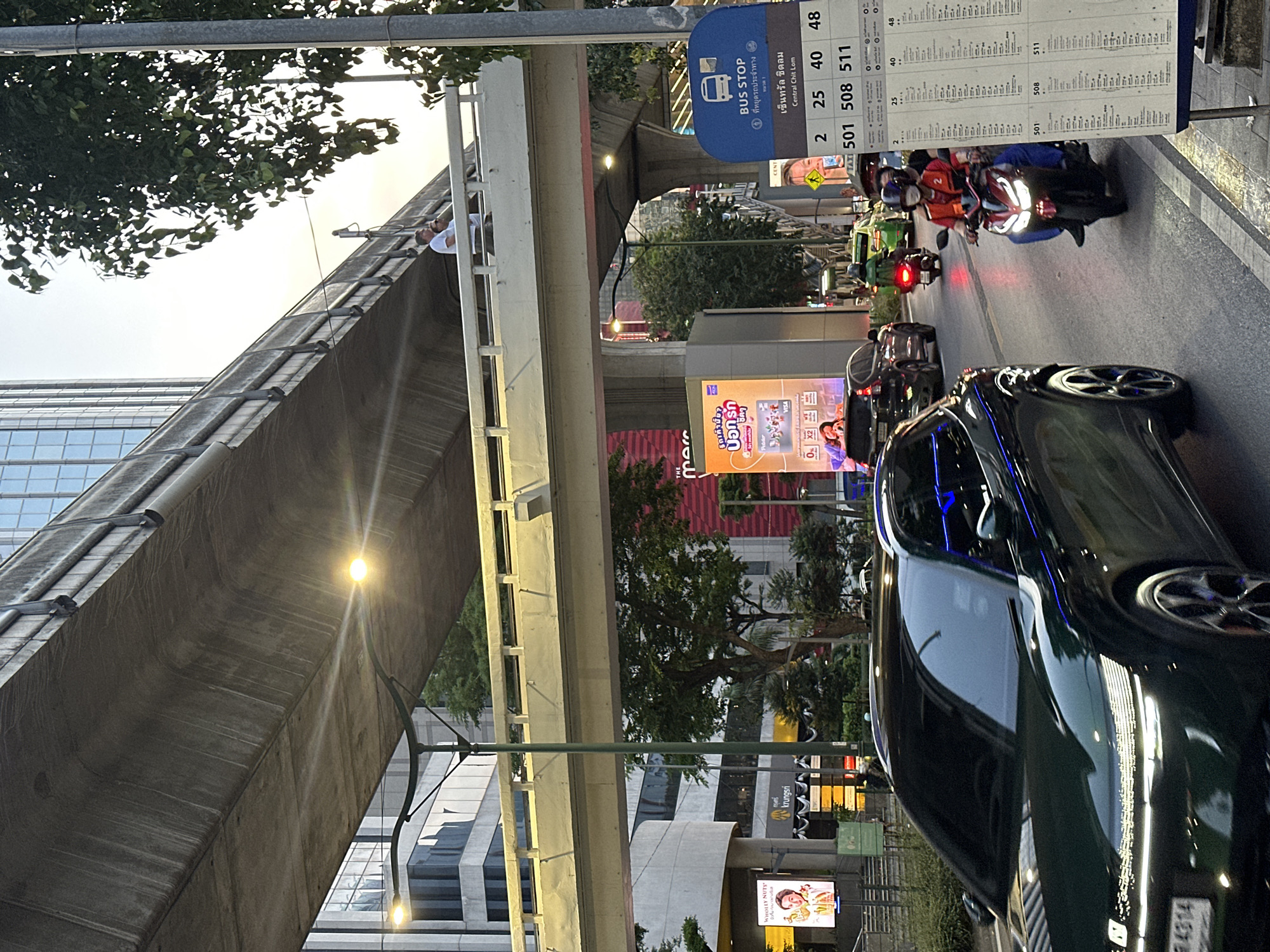 The image captures an urban scene in Bangkok, Thailand, likely in the Central Chit Lom area. The presence of a bus stop sign with the designation "Central Chit Lom" reinforces this.
Vehicles on the road include a dark-colored sedan, several motorcycles with riders, and a green taxi. Advertisements and signage are visible, including a "Bounty Nutty" advertisement, the name "krungsri," and the Thai text "ยกเค้ายา บอกรัก".
An elevated structure, possibly a pedestrian walkway or part of the BTS Skytrain system, spans the road. Two people are visible on the walkway.
The overall environment is a busy city street with trees and buildings in the background.