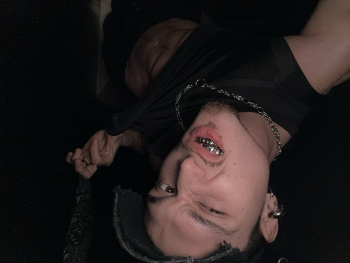 The image features a young adult male positioned in a dimly lit, dark environment. The subject has light skin, dark hair, and a short beard/goatee. He wears a distressed, dark baseball cap with a torn brim and multiple silver ear piercings/cuffs on his left ear. A prominent silver chain necklace is visible around his neck. His mouth is open, revealing a custom silver grill covering both his upper and lower teeth. His face is contorted in a grimace or a playful, intense expression, with his right eye visible and squinted. He is wearing a dark sleeveless top or vest, which he is actively pulling up with his right hand, exposing his abdomen and defined abdominal muscles. The background is uniformly dark and devoid of specific identifiable features. The exact location within the city of Sofia, Bulgaria, cannot be determined from the visual information provided in the image.