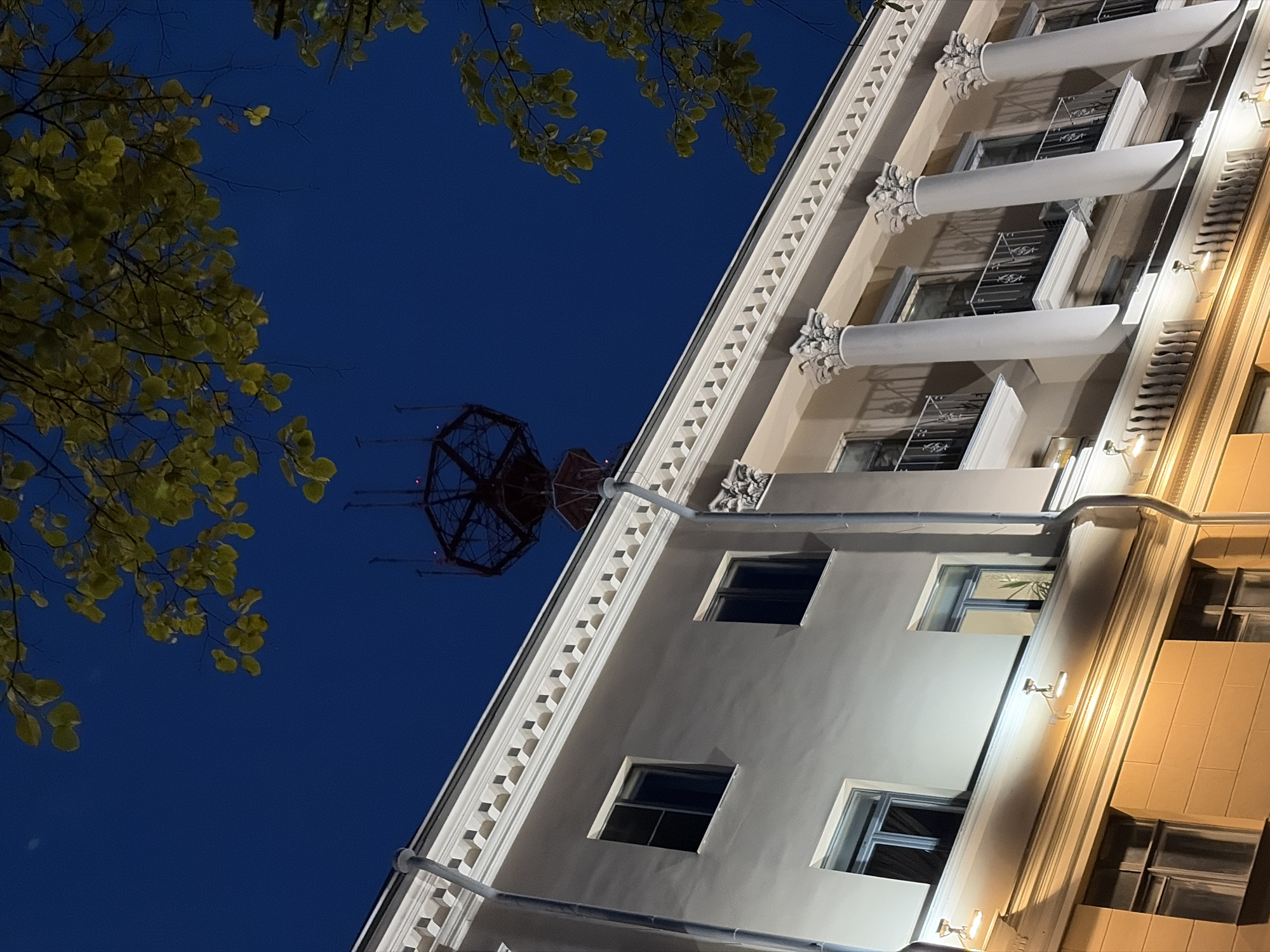 The image shows the top floors of an old building at night, with a dark blue sky and some illuminated branches of a tree in the foreground. The building has a cream-colored facade with white decorative elements, including a cornice with geometric patterns and large columns topped with ornate capitals. There are several windows, some with balconies and black metal railings. The lower part of the building, visible at the bottom, is illuminated by warm yellow lights, suggesting it might be a different level or a separate structure. A distinctive, dark, lattice-like tower structure with red lights is visible against the sky, positioned above and behind the building. This tower resembles an antenna or transmission tower. The overall impression is of an urban scene at dusk or night, with architectural elements characteristic of European cities. The presence of the unique tower structure and the architectural style are indicative of the building located at **Independence Avenue, 103, Minsk, Belarus**, which is the former KGB headquarters and currently houses the Ministry of Defense and the General Staff of the Armed Forces of Belarus.