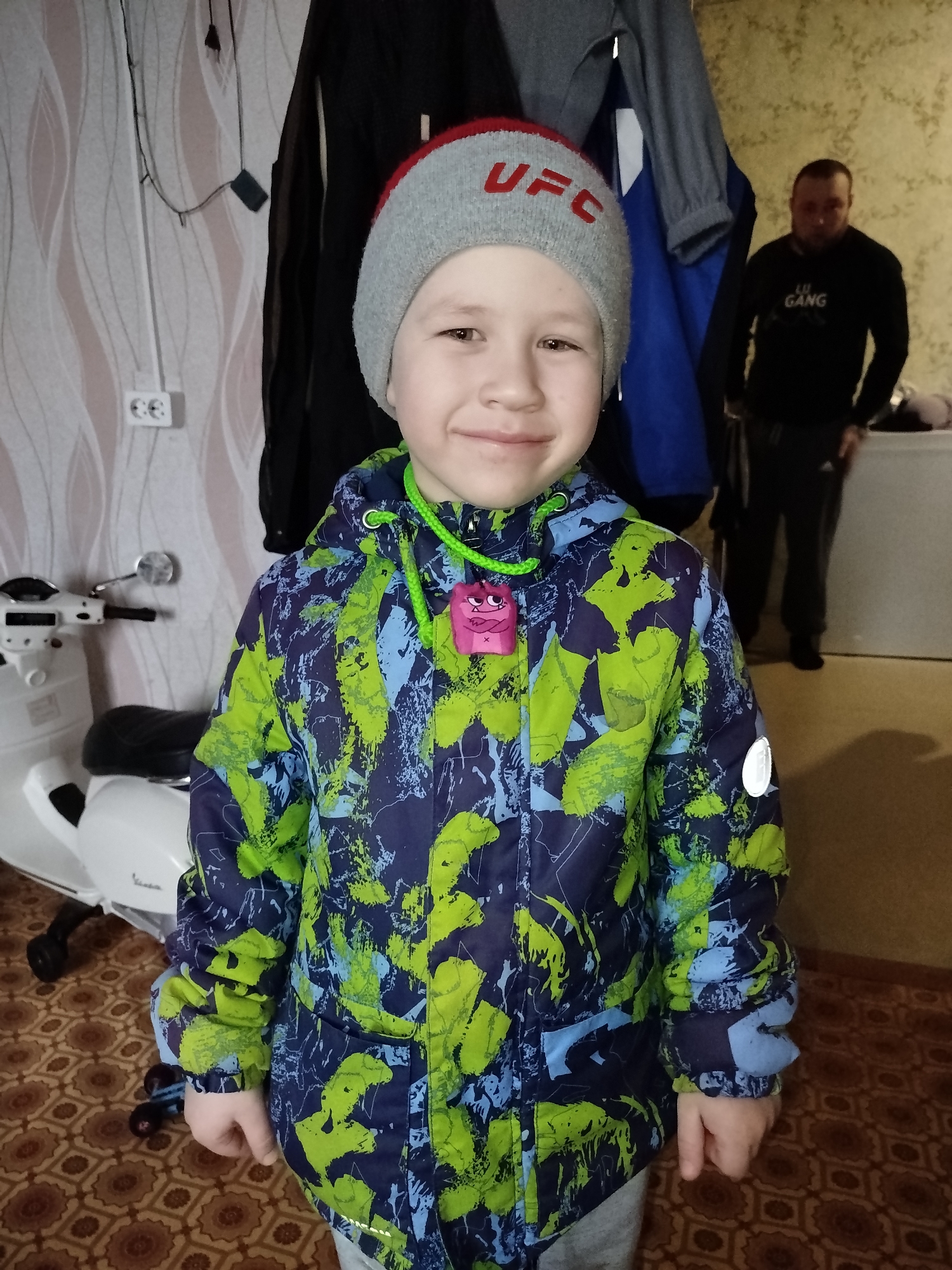 The image shows a young boy standing indoors. He is wearing a grey and red "UFC" hat and a colorful blue, green, and navy patterned jacket with a green drawstring and a pink monster-shaped pendant. He is smiling. Behind him to the left is a white scooter. To the right, a man stands in the doorway wearing a black sweatshirt with the words "LU GANG" on it and dark-colored pants. Clothes hang on a rack behind the boy. The background wall has a patterned wallpaper, and there is a brown patterned rug on the floor. The location is likely Chaikovsky, Russia.