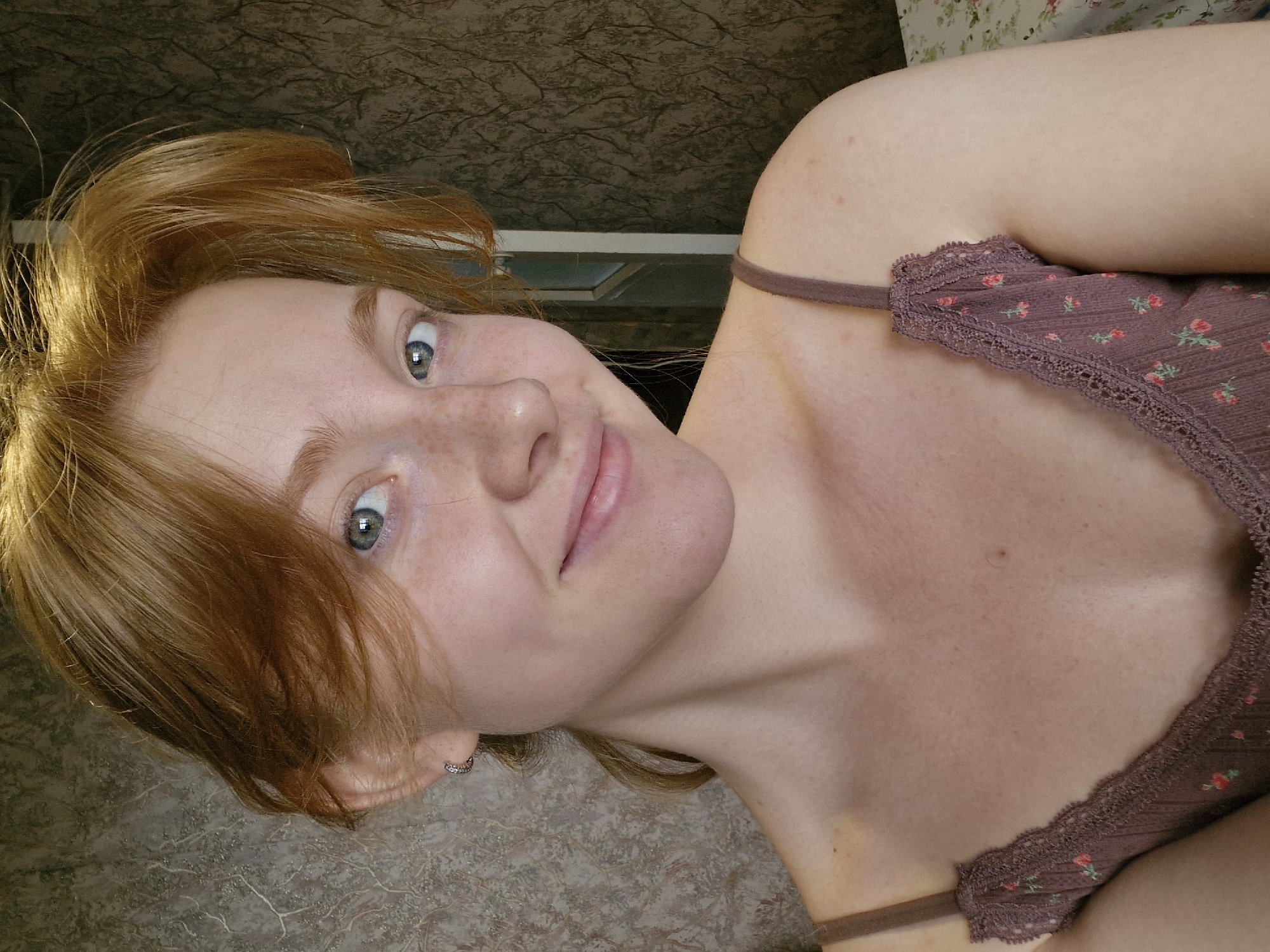 A close-up, angled shot features a fair-skinned young woman with red-blonde hair and freckles. Her head is tilted, and she is looking directly at the viewer with a slight smile. She is wearing a brown camisole top with a floral pattern and lace trim. The background consists of a patterned wall and a partial view of what appears to be a window frame. The city and location are unknown.