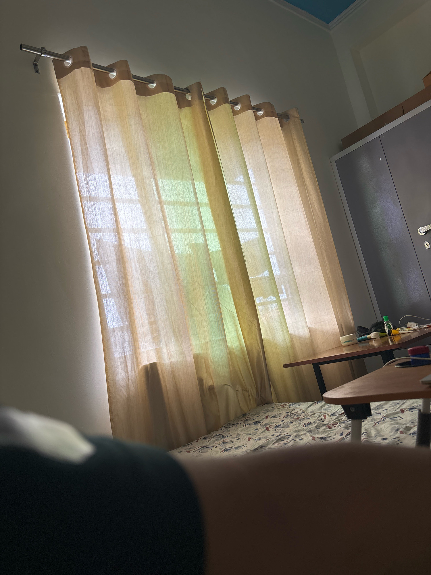 The image depicts an indoor scene, likely a bedroom, with natural light filtering through a window covered by sheer, light-colored curtains. The curtains hang from a silver curtain rod mounted on the wall. The window itself is visible as a grid pattern through the fabric.
To the right of the curtains, a gray metal wardrobe or cabinet stands against the wall. On a wooden table in front of the wardrobe, several items are placed, including a small green bottle, a white container, some pens or markers, and a black bag. A second, lower wooden table is partially visible in the foreground, also on the right side of the image. The floor is covered with a patterned bedsheet, suggesting a bed is present, though it is mostly obscured.
In the lower-left corner of the image, a portion of a person is visible, appearing out of focus and dark, possibly a shoulder or head covered in dark clothing. The overall lighting suggests it is daytime, with sunlight creating bright areas and shadows. The ceiling is painted blue and white. The location is specified as Bhatta, India.