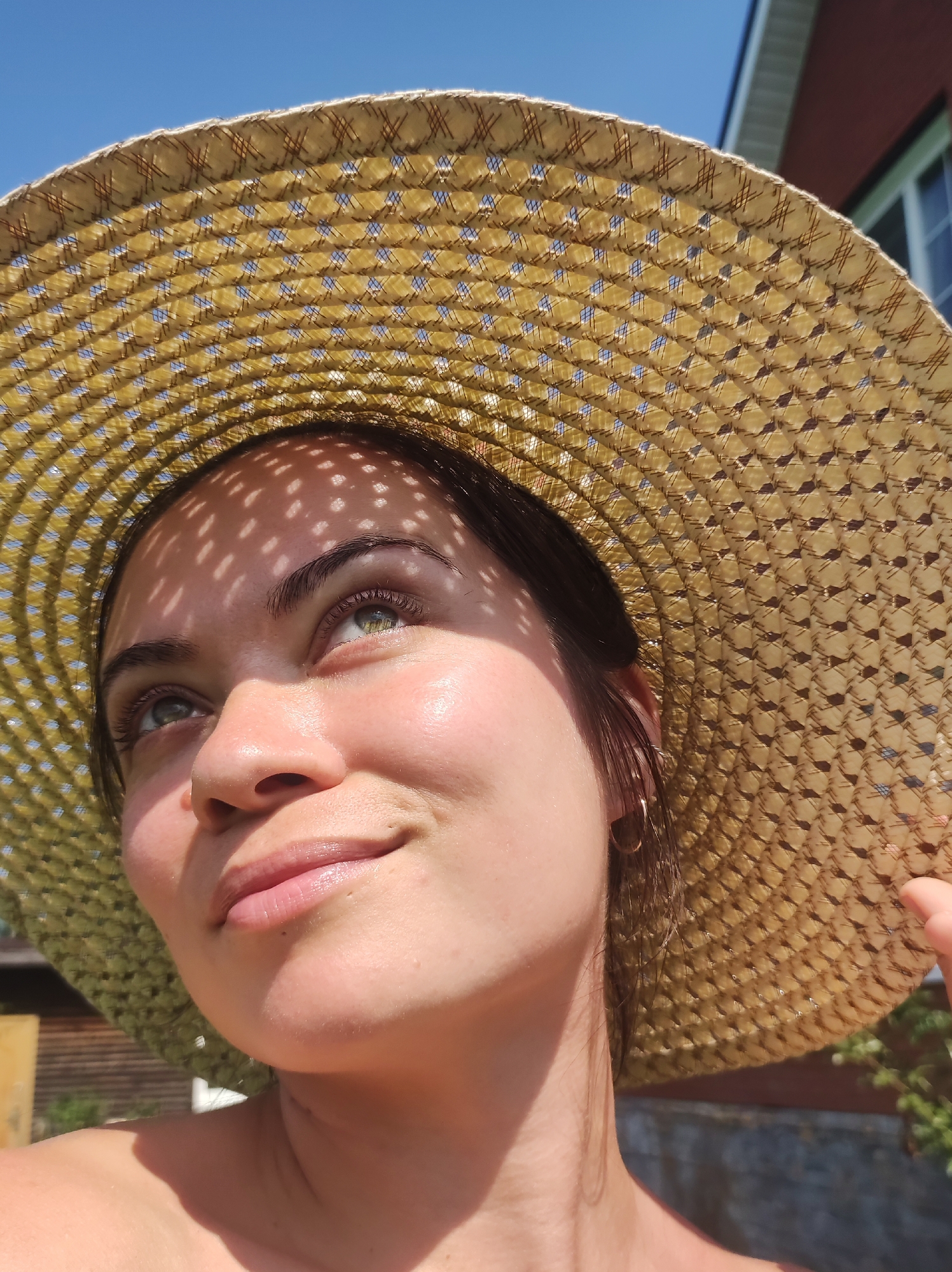 The image shows a close-up, low-angle shot of a young woman wearing a wide-brimmed, woven straw hat. The hat casts dappled shadows on her face, highlighting her upward gaze. She has light skin, dark hair, and appears to be looking towards a bright light source. The background is a clear blue sky with a hint of a building and foliage. The scene suggests an outdoor setting on a sunny day, possibly in a suburban or rural area given the presence of the building and greenery. There is no indication of the specific location within the city of Фоки, Russia.