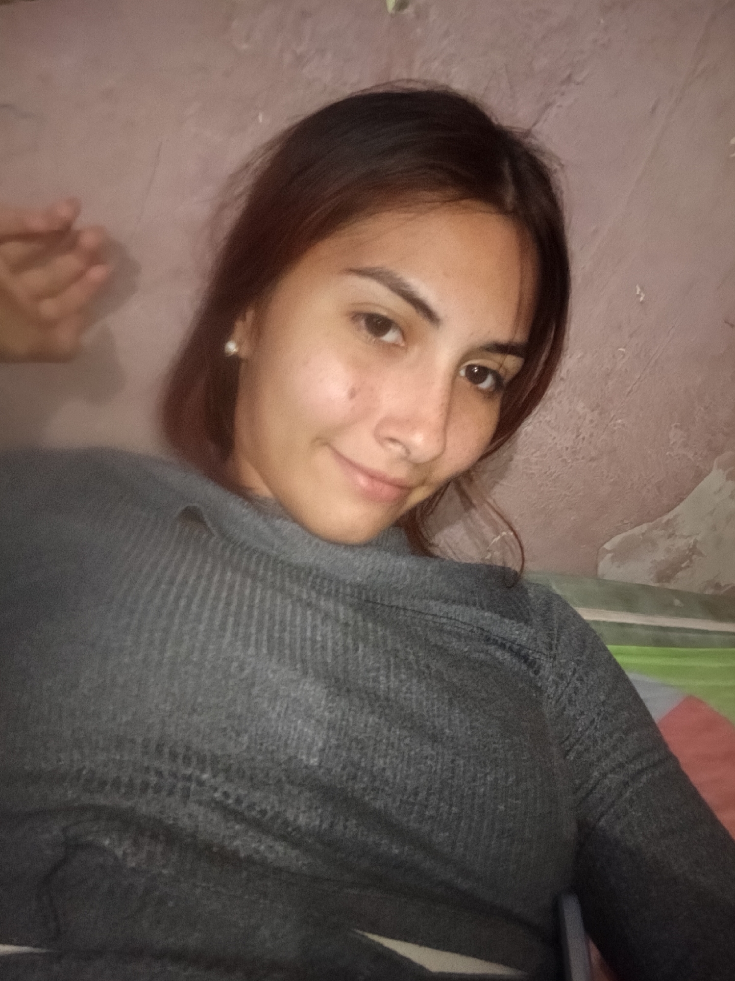 A young woman with shoulder-length brown hair and fair skin is shown in a close-up selfie. She is wearing a gray sweater. The background is a textured, light pink wall, and there is a disembodied hand slightly out of focus in the upper left corner. The location of Juan Griego, Venezuela is not verifiable from this image.