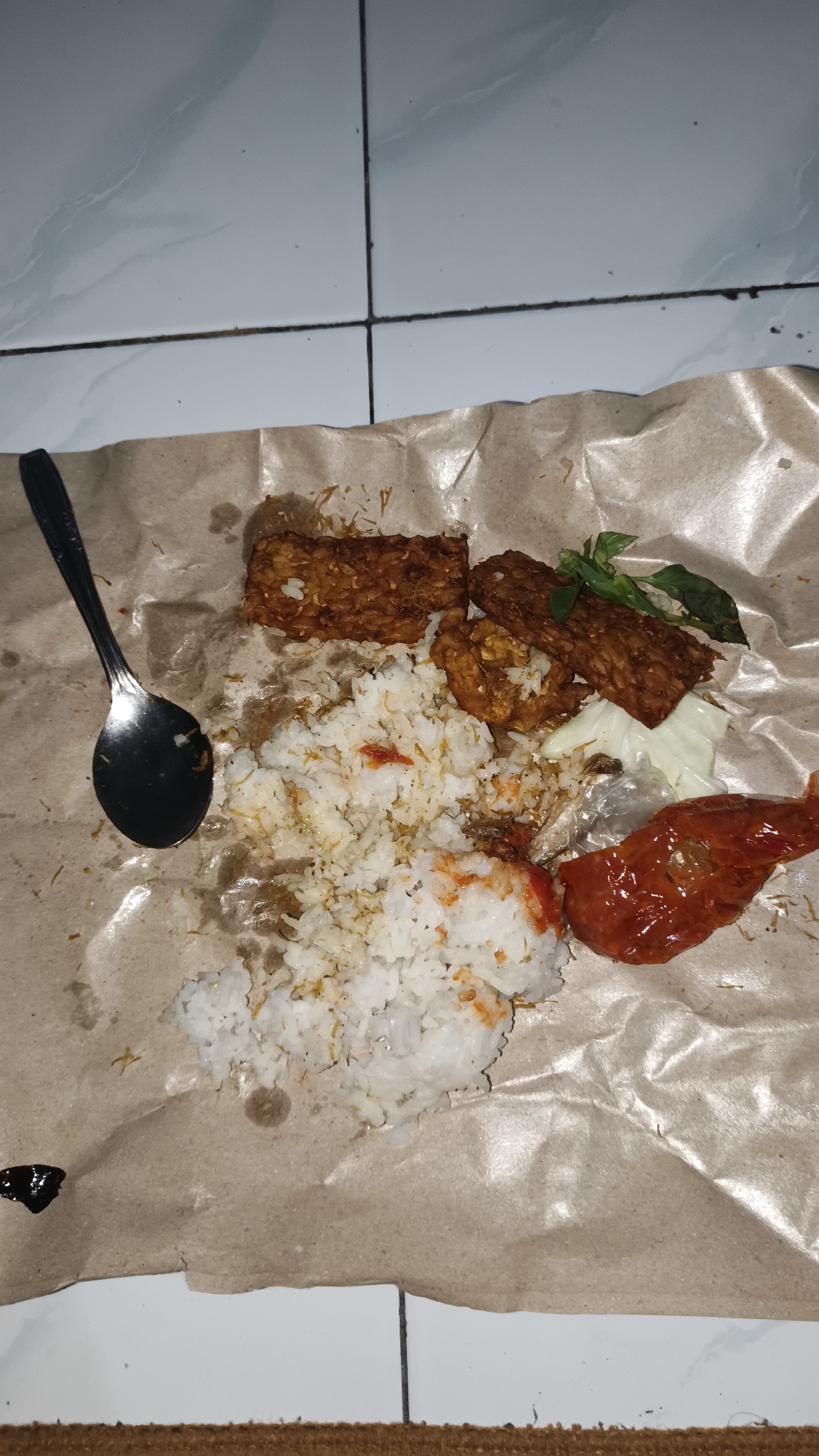 A close-up, overhead shot displays a plate of food served on brown paper. The food consists of white rice, several pieces of fried tempeh, and a piece of red chili sauce. A metal spoon rests to the left of the rice. The background shows a tiled floor and a brown mat. The scene appears to be indoors. There is no visible text.