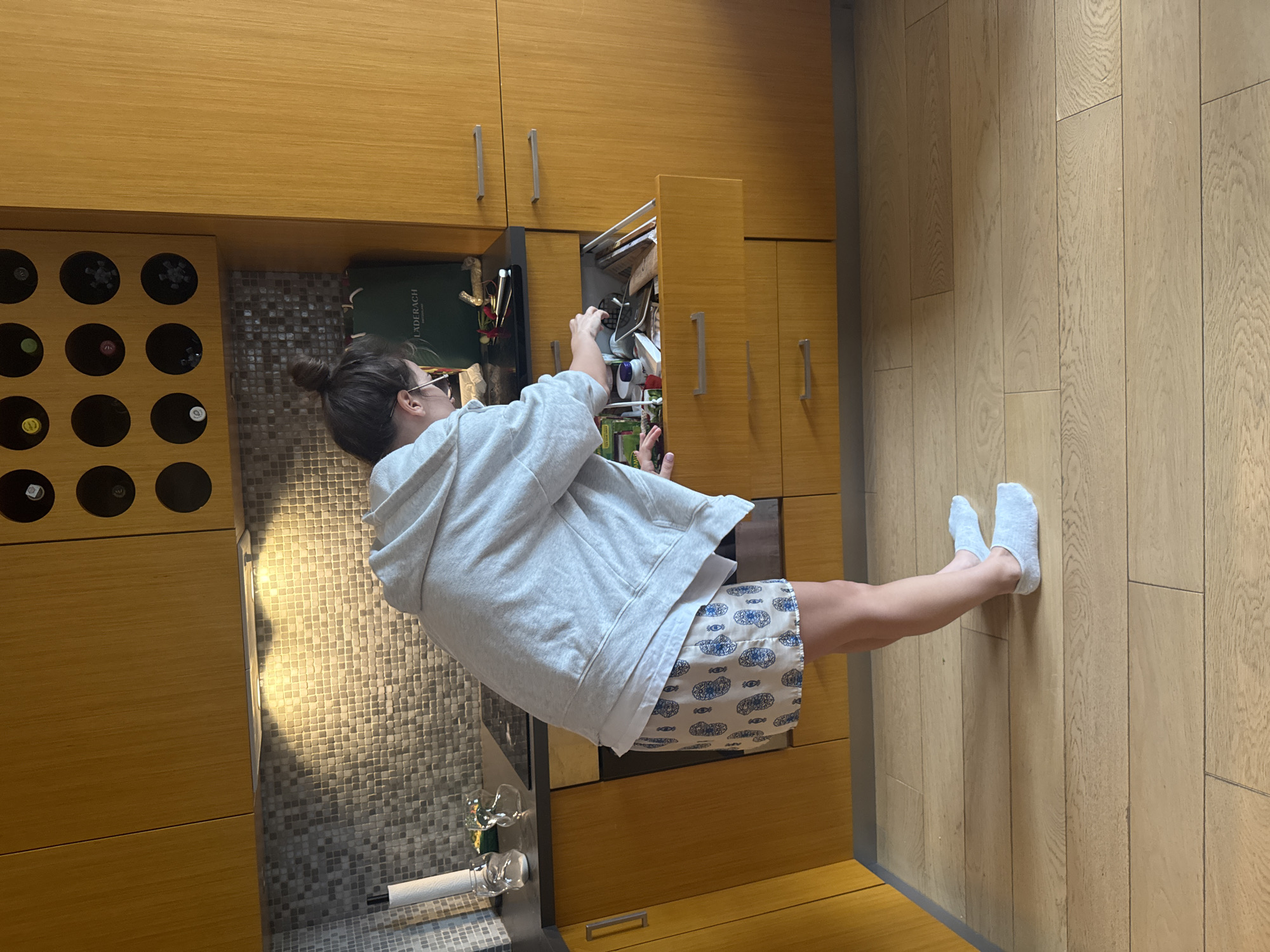 A person is shown from a slightly elevated, side-back perspective, standing in a modern kitchen or pantry and reaching into an open pull-out drawer or cabinet. The image appears to be rotated, with the person standing upright relative to the floorboards.
The subject is a female with her hair tied in a bun, wearing glasses, a light grey hooded sweatshirt, and patterned blue and white shorts. She has light blue socks on her feet. Her hands are inside the open drawer, which is filled with various items, suggesting she is searching for or retrieving something.
The setting features light wooden cabinetry with silver bar handles. To the left, an integrated wooden wine rack holds several bottles. Below the upper cabinets and behind the person, there is a mosaic-tiled backsplash in shades of grey. A dark countertop is visible, on which a green bag with the text "LADERACH" is placed, along with what appears to be a glass and a paper towel roll. The floor consists of light wooden planks. The area is well-lit, with light reflecting off the backsplash.