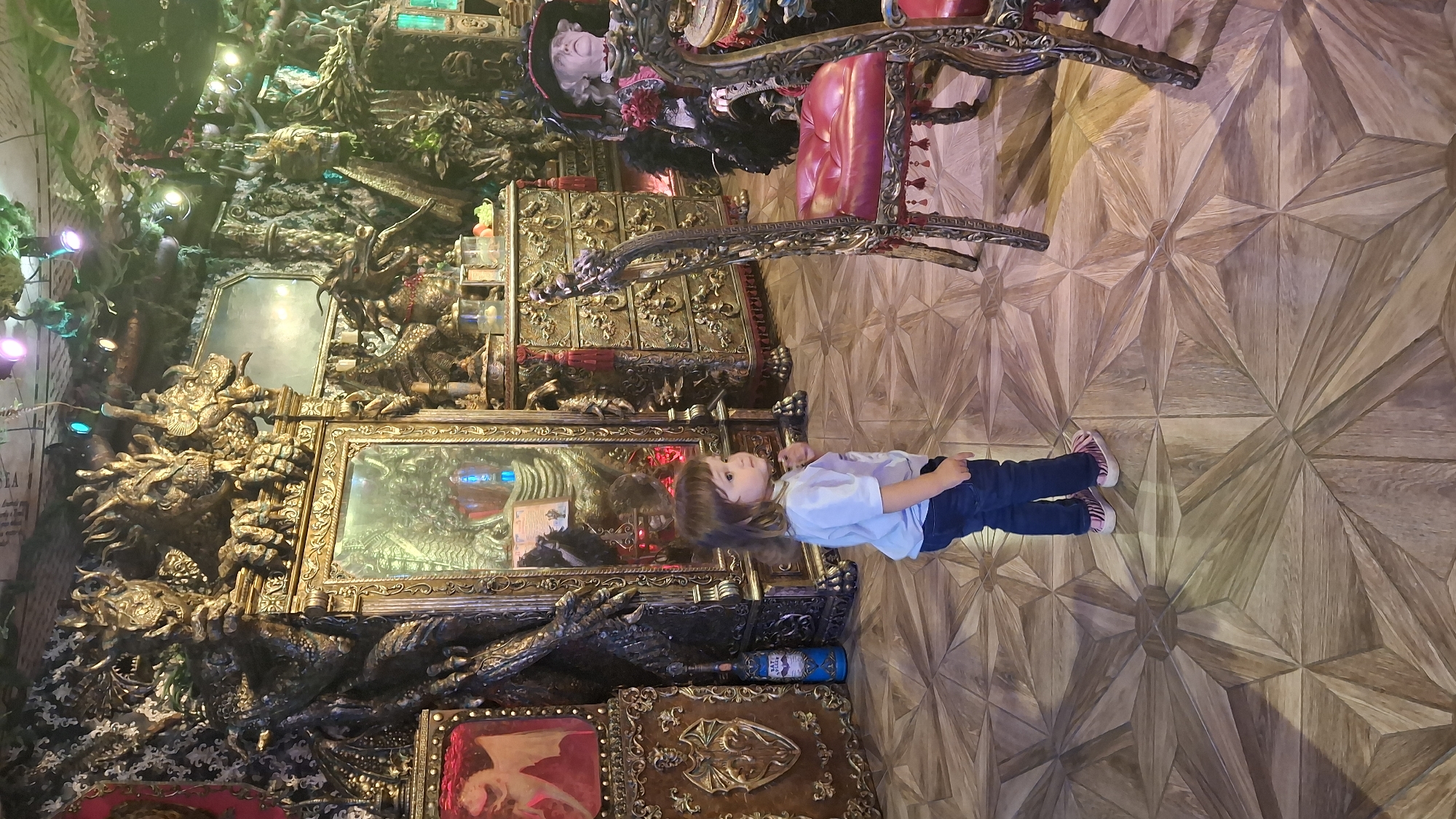 The image depicts a young child standing in a room adorned with elaborate, gothic-style decor. The room features a large, ornate mirror framed by dragon-like sculptures, gilded furniture, and a dramatic, dark aesthetic. The floor is covered in a patterned wooden parquet. In the background, a mannequin or bust dressed in a historical, theatrical costume is visible. The overall impression is one of a unique, possibly themed, interior. The scene is set within the "Big Book Cafe" located at 32 Nevsky Prospect in Saint Petersburg, Russia.