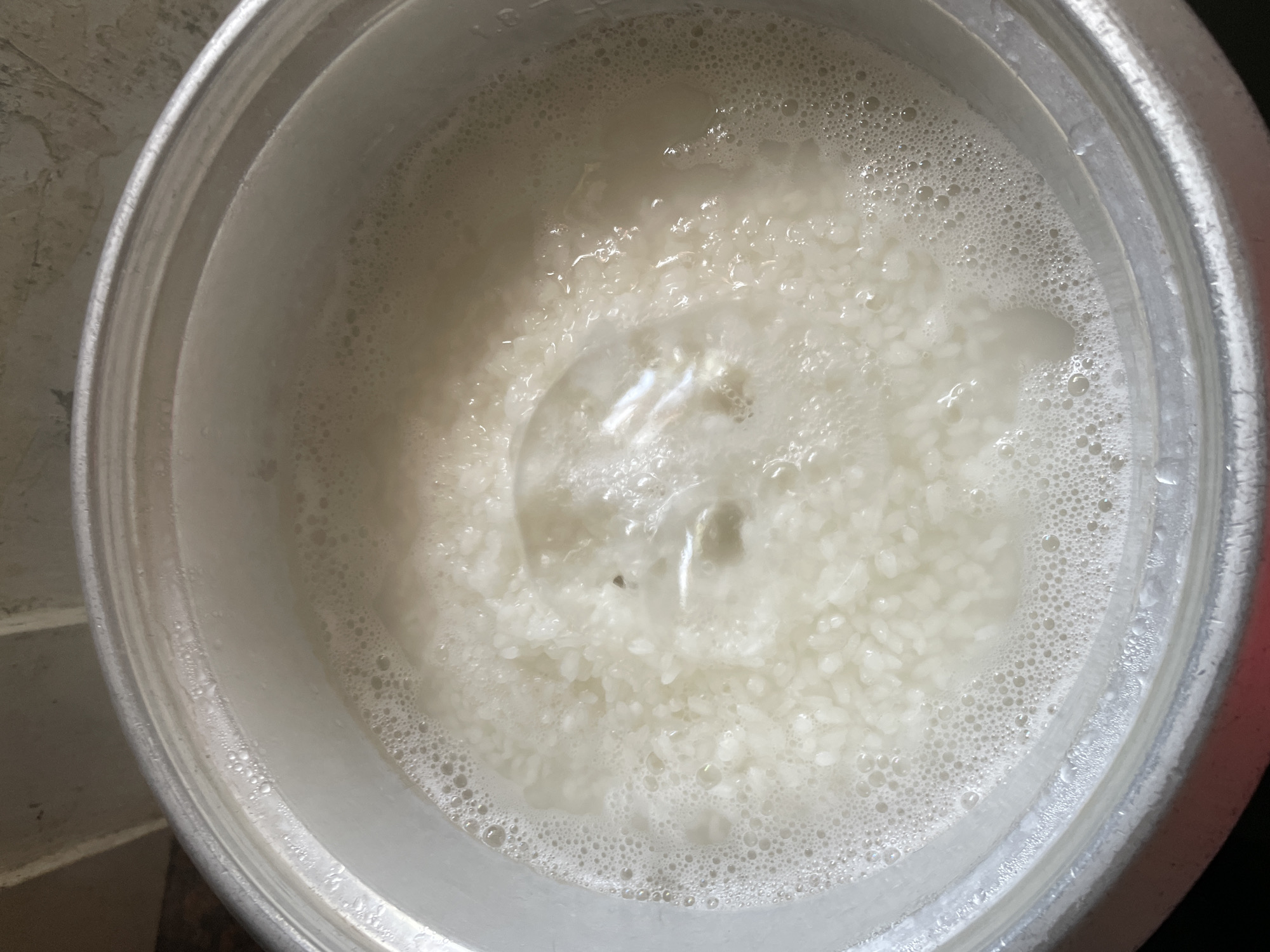 An overhead shot captures the interior of a metallic pot, likely a rice cooker insert, filled with white rice actively boiling in water. The scene suggests a domestic setting, possibly a kitchen, in Davao City, Philippines, where a meal is being prepared.
The pot's interior is predominantly filled with milky-white water, thick with foam and small bubbles rising to the surface, indicating vigorous boiling. Numerous individual white rice grains are visible beneath the frothy surface, especially towards the bottom and sides of the pot. A larger, more turbulent bubble or disturbance is noticeable in the center of the liquid.
The rim of the metallic pot, which appears to be silver-colored, is visible around the perimeter of the image. Faint markings, such as "1.0" followed by other indistinct characters (likely indicating a volume measurement), can be seen etched or printed on the upper left interior edge of the pot. The exterior of the pot and the surrounding environment are mostly out of focus or cropped out, but hints of a light-colored, possibly textured surface are visible near the outer rim. The overall impression is one of simple, everyday food preparation.