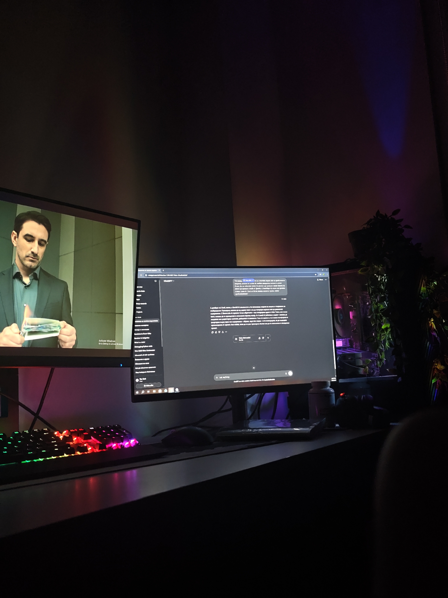 A dimly lit indoor scene shows a desk with two computer monitors. The monitor on the left displays a man on screen holding burning money. The monitor on the right shows an open ChatGPT interface. A colorful gaming keyboard and a mouse sit on the desk. A desktop computer case with internal lights sits to the right of the monitor and is partially obscured by plants. Bottles are also visible on the desk. The walls are dark, with a blue light shining in the corner. No location in Kozloduy, Bulgaria, is evident.