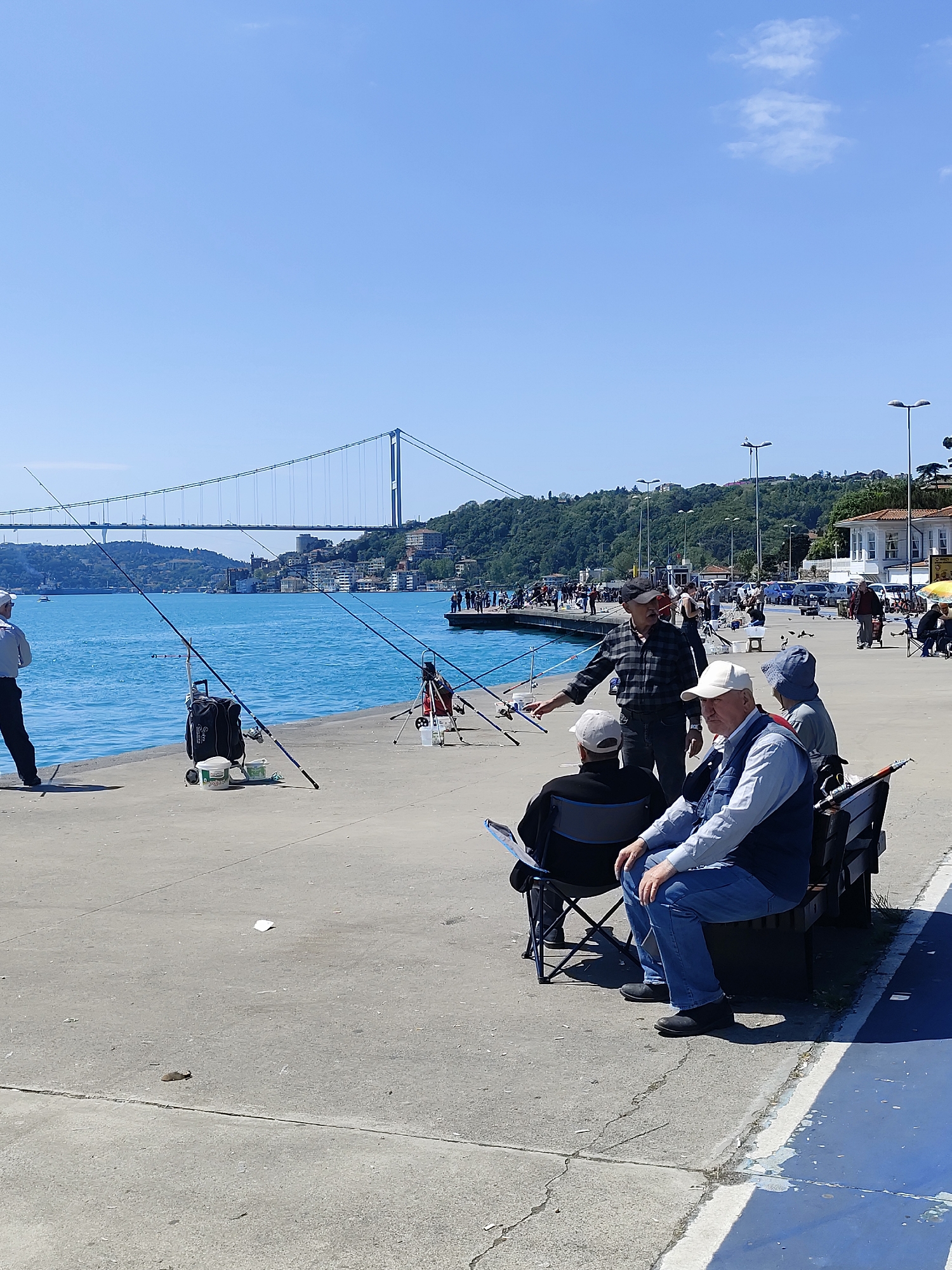 The image depicts a waterfront scene in Sarıyer, Turkey, with the Bosphorus Bridge visible in the background. Several individuals are engaged in fishing activities along the concrete pier. Fishing rods are set up, some with accompanying gear like tackle boxes. A group of men are seated on a bench, while another man in a checkered shirt stands nearby, gesturing towards the fishing lines. The sky is clear and blue, and the water is a turquoise color. People are walking along the pier further in the background. Streetlights line the edge of the pier.