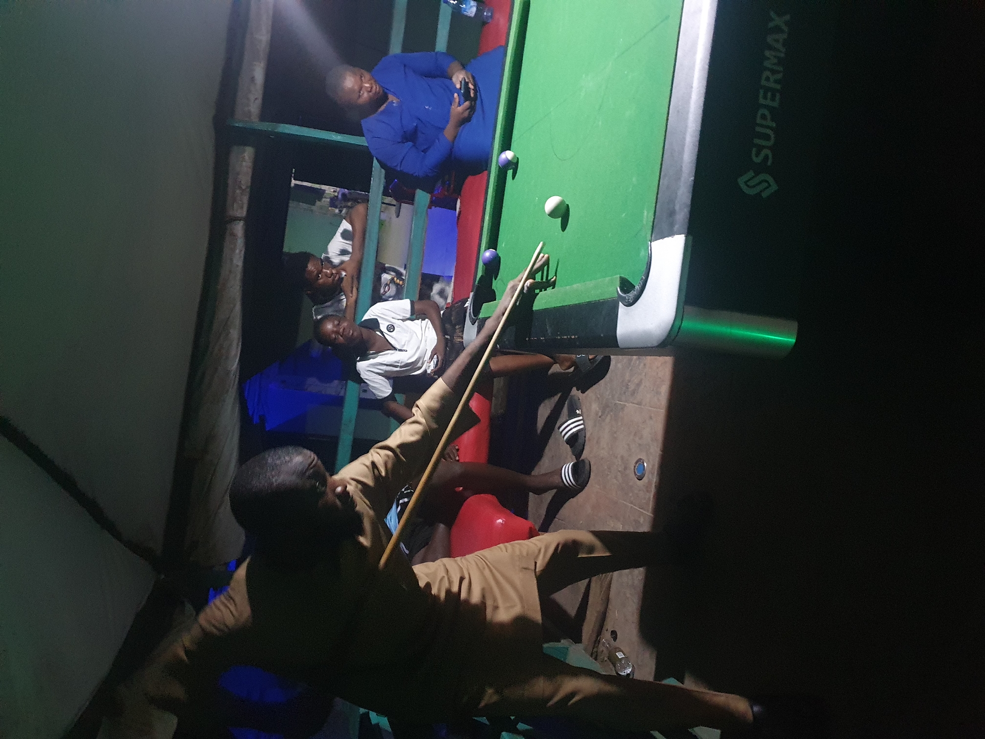 In Lokoja, Nigeria, a group of individuals is gathered around a pool table under a canopy. A man, dressed in a tan-colored outfit, is actively playing pool, positioned with a cue stick in his hands, aiming at the balls on the green felt. The pool table, a "Supermax" brand, is well-lit, contrasting with the dim surroundings. Three other people are seated or standing in the background, observing the game. One woman, wearing a blue outfit, is seated and appears to be using her phone. Another individual, a young man in a white shirt, is leaning against a railing, also watching. A third person, also appearing to be a young man, is seated to the left, wearing a white shirt. The environment suggests an outdoor recreational space, possibly a bar or a casual gathering spot, with tiled flooring and some debris on the ground, including a discarded plastic bottle. The lighting is dim, with the primary light source illuminating the pool table and the players.