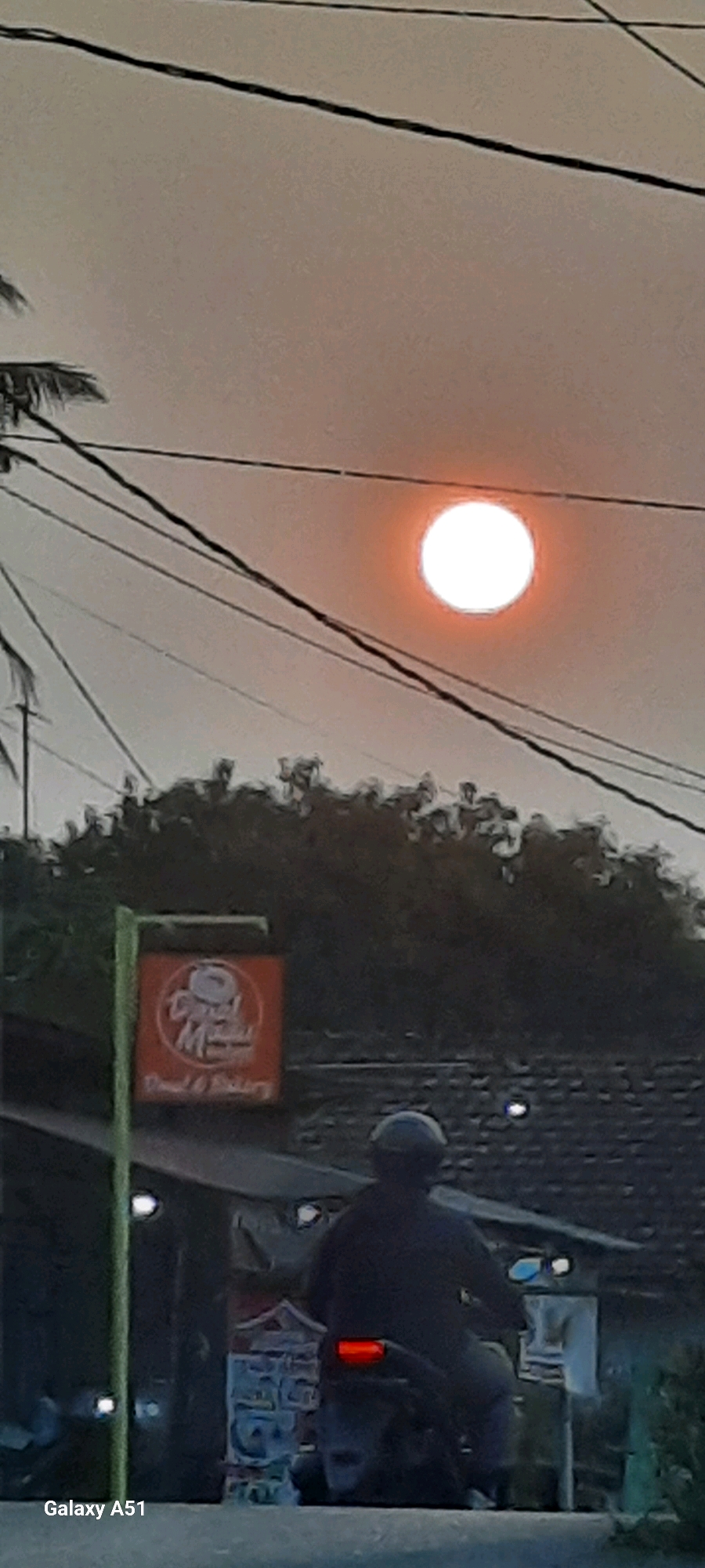 The image depicts a street scene in Selopuro, Indonesia, during what appears to be sunset or sunrise. A bright, orange-tinted sun is visible in the sky, partially obscured by power lines. In the foreground, a person on a motorcycle is silhouetted against the light. To the left, a red sign with a donut graphic and text that reads "Mamad" and "Donut & Bakery" is mounted on a green pole. The background consists of trees and the roof of a building, suggesting a semi-urban or rural setting. The overall lighting is dim, with the sun being the primary light source. The photograph is taken from a slightly low angle, looking upwards towards the sky and the subjects.