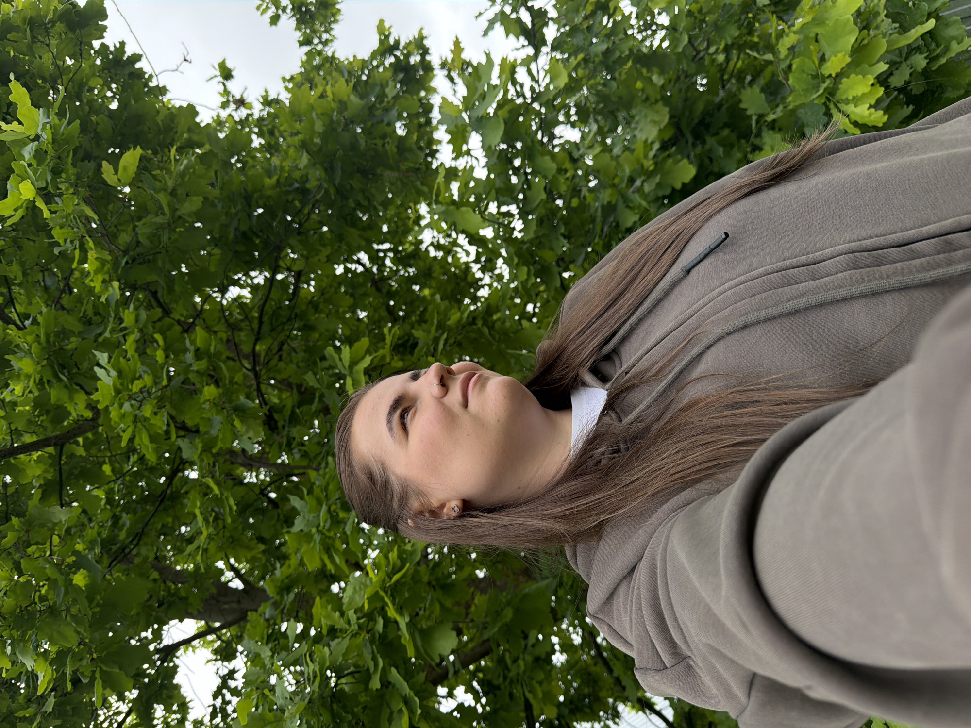 A young woman with long, straight brown hair is captured in a medium shot, looking upwards and slightly to her left with a serene expression. She is wearing a plain grey or khaki-colored hooded sweatshirt, beneath which a white collar is visible. A small silver ring adorns her left nostril, and a subtle earring can be seen in her left earlobe.

The scene is set outdoors amidst a lush green tree canopy, predominantly featuring broad, lobed leaves characteristic of oak trees. The dense foliage creates a vibrant backdrop, with bright, overcast sky visible through the gaps in the leaves, indicating daytime, likely during late morning or afternoon. The upward-looking perspective suggests the photograph was taken from a lower angle, possibly while the woman was standing or sitting beneath the trees. The overall mood is calm and natural.

The image suggests an outdoor portrait in a natural setting like a park or forest, in Marakushi, Russia. There is no visible text in the image.