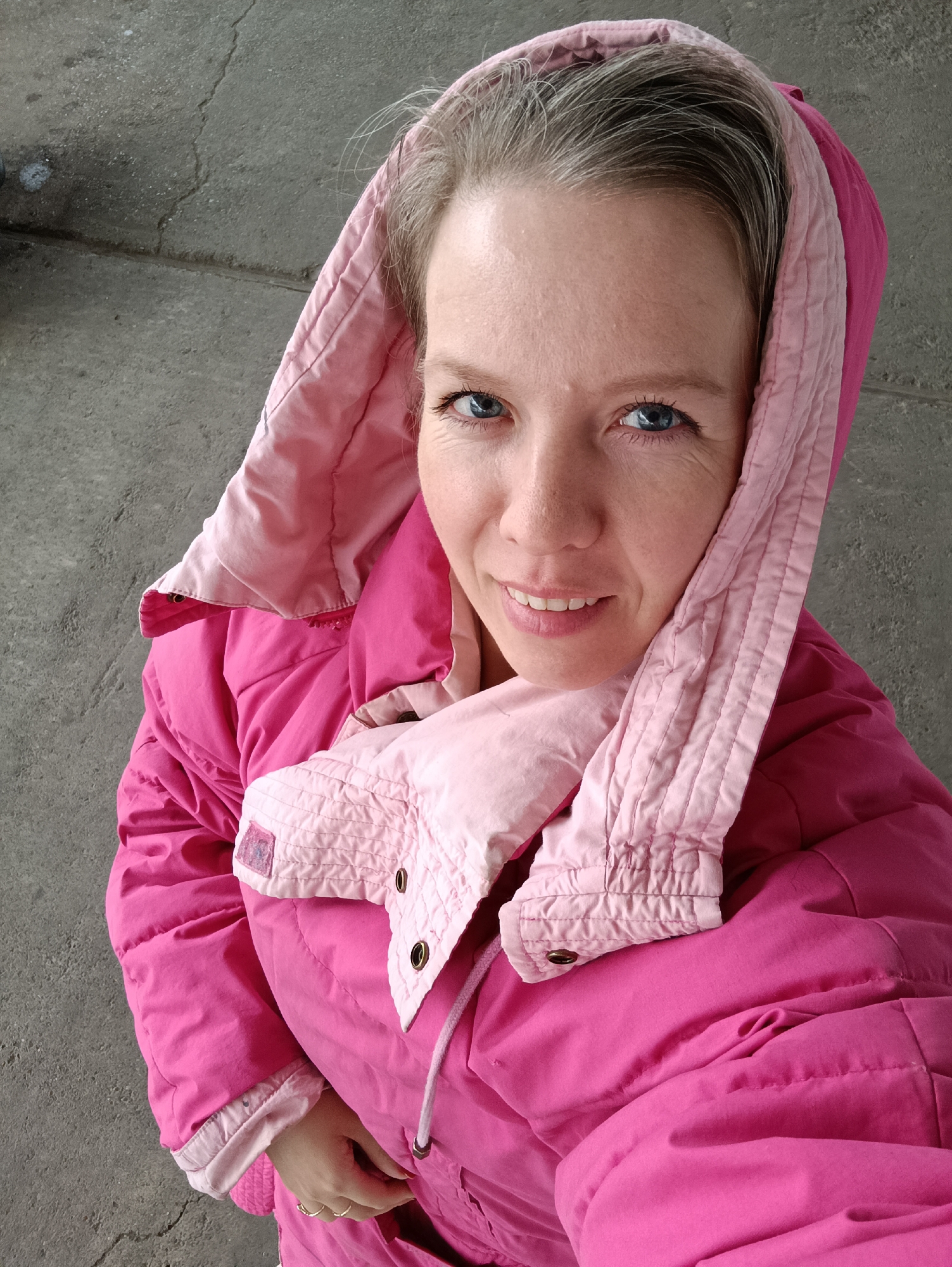 A woman is posing for a photo outdoors. She is wearing a pink hooded jacket. The background appears to be a concrete surface, possibly a sidewalk. The location and city cannot be determined from this image alone.