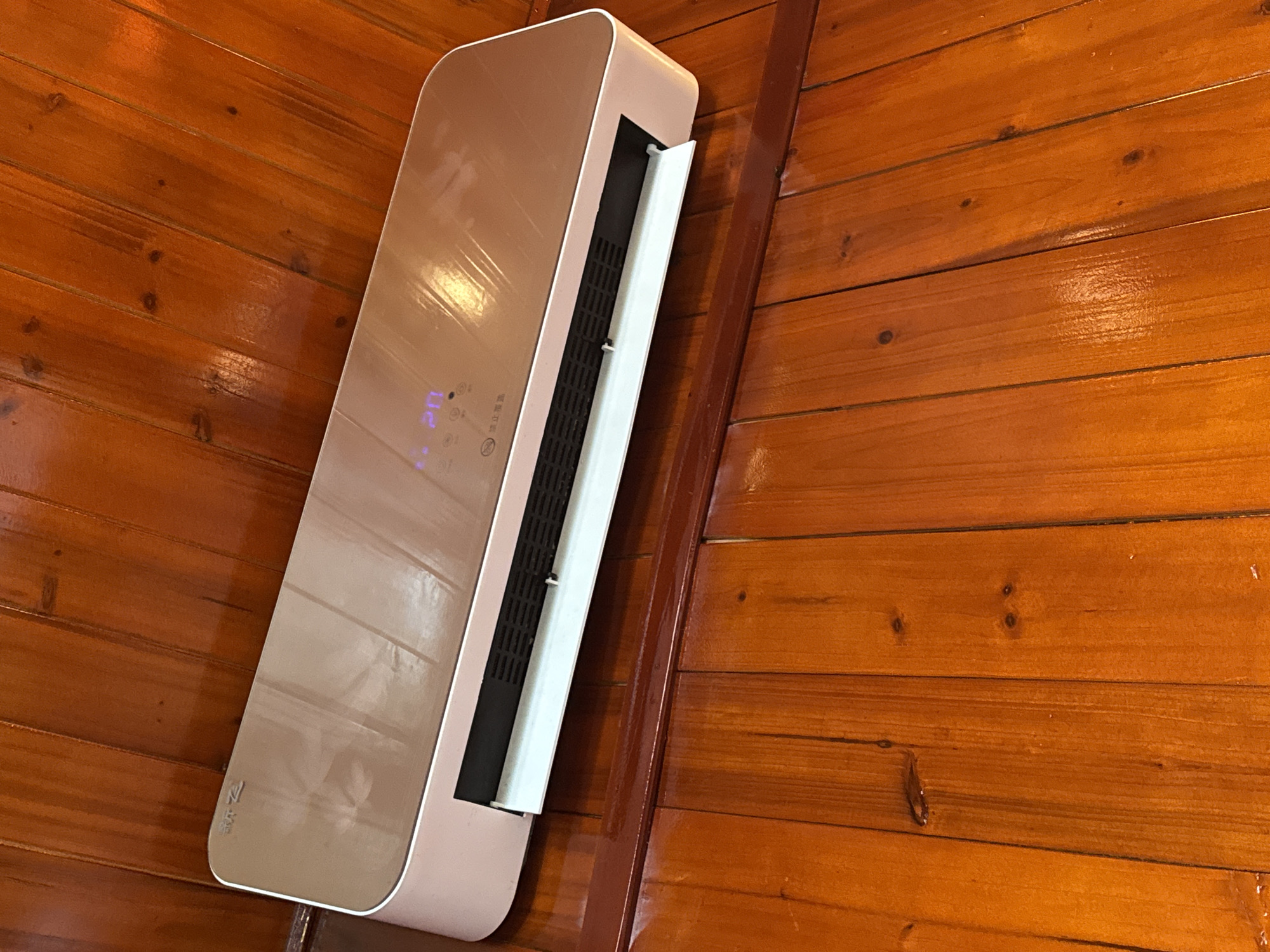 The image shows a wall-mounted air conditioning unit in a room with wood-paneled walls. The air conditioner is a light brown and white color, with an open vent revealing a black filter inside. The wood paneling consists of horizontal planks with a glossy finish. There is nothing to specify the location of this place is in Binh Lu, Vietnam.