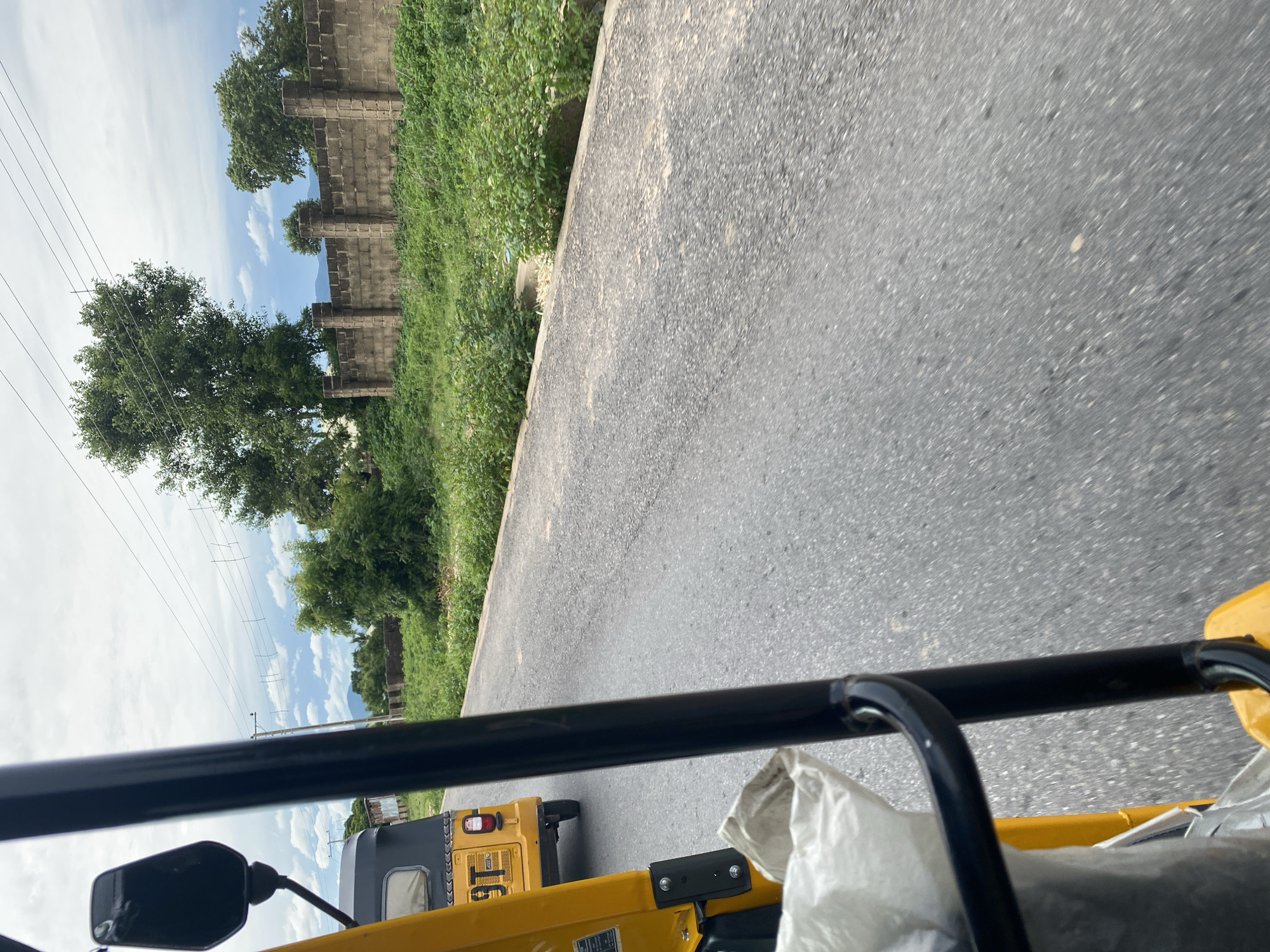 The image depicts a view from a yellow vehicle, likely an auto-rickshaw, traveling on a paved road. The perspective is from inside the vehicle, looking forward and slightly to the side. In the foreground, the black frame of the vehicle's roof and a side mirror are visible.
To the left of the road, there is a grassy area with lush green vegetation and several trees. Beyond the vegetation, a brick wall with decorative pillars runs along the scene, suggesting a boundary or property line. The sky above is a mix of blue and white clouds, indicating daytime with fair weather. Power lines are also visible stretching across the sky, supported by poles.
Further in the distance, more greenery and some structures are faintly visible. The road itself is asphalt and appears to be in good condition. Another yellow auto-rickshaw is seen ahead, also moving along the road.
The image's orientation is rotated, making the scene appear sideways. The overall impression is of a journey on a sunny day in a semi-urban or rural area, possibly in Jalingo, Nigeria, as indicated by the location context. No people are clearly visible, but the presence of the vehicles implies human activity. There is some visible text on the yellow vehicle, but it is not legible in this image.