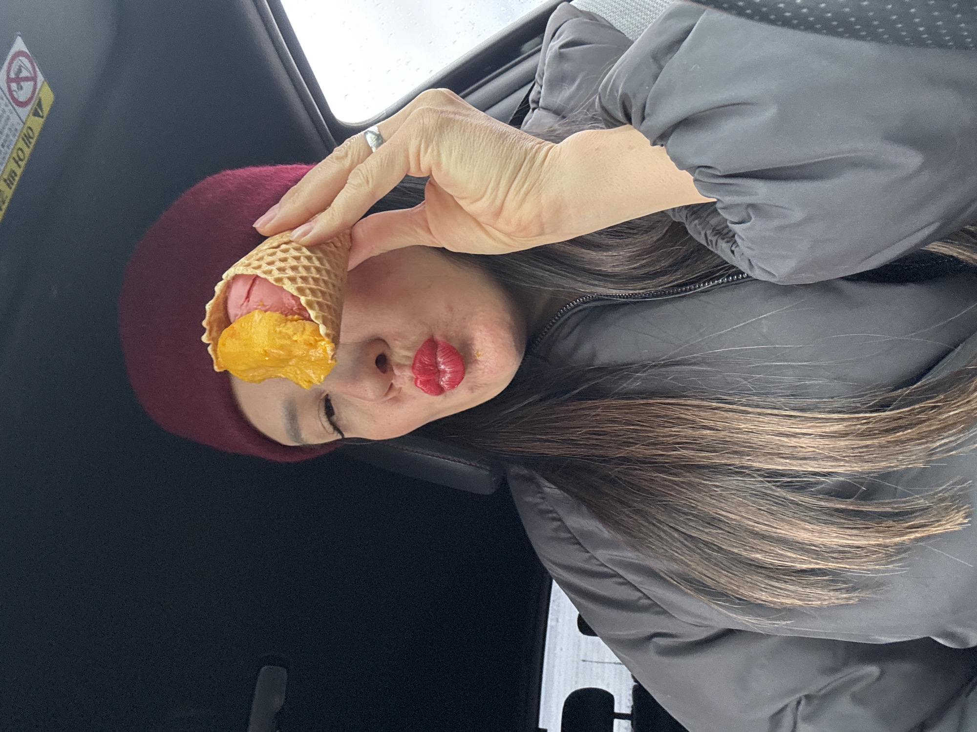 The image depicts a person inside a car. The person is wearing a burgundy hat and a grey jacket. They are holding an ice cream cone with two scoops: one pink and one orange. The person's lips are bright red. The background shows the car's interior, including the black door panel with a yellow warning sticker.
There is no information in the image to determine the location within Chaikovsky, Russia, or if it is even in that city.