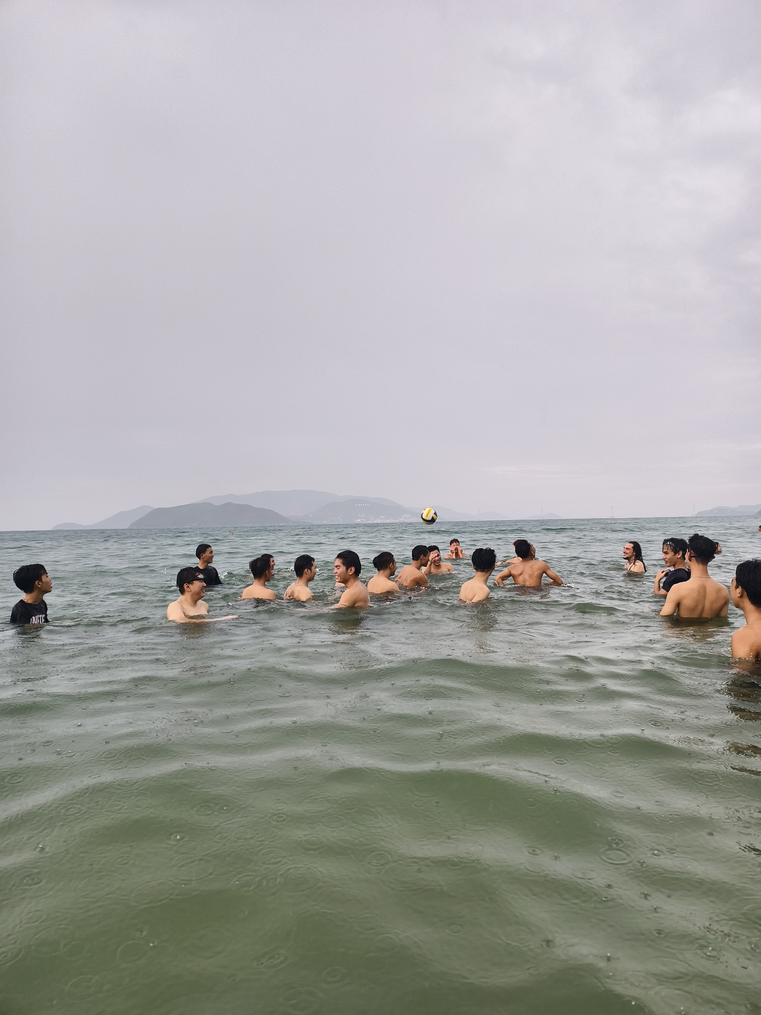 A group of approximately 15 to 20 young men are gathered in a large body of water, likely the sea or a sizable lake, under an overcast sky. Most are shirtless, with their heads and shoulders visible above the water. One person on the far left wears a dark t-shirt with the word "IGNITE" partially visible on it.
The men appear to be engaged in a game of water volleyball, as a yellow and white striped volleyball is suspended in the air or floating on the water in the mid-ground. Several individuals are looking towards the ball, while others are conversing amongst themselves, exhibiting engaged or smiling expressions.
The weather is cloudy and overcast, contributing to a muted lighting. Raindrops are clearly visible on the surface of the water, indicating that it is raining lightly or has just rained. In the distance, a hazy outline of mountains or landmasses stretches across the horizon. The water has a greenish-grey hue. The overall scene depicts a casual group recreational activity in the water during daytime, despite the cloudy and rainy conditions.