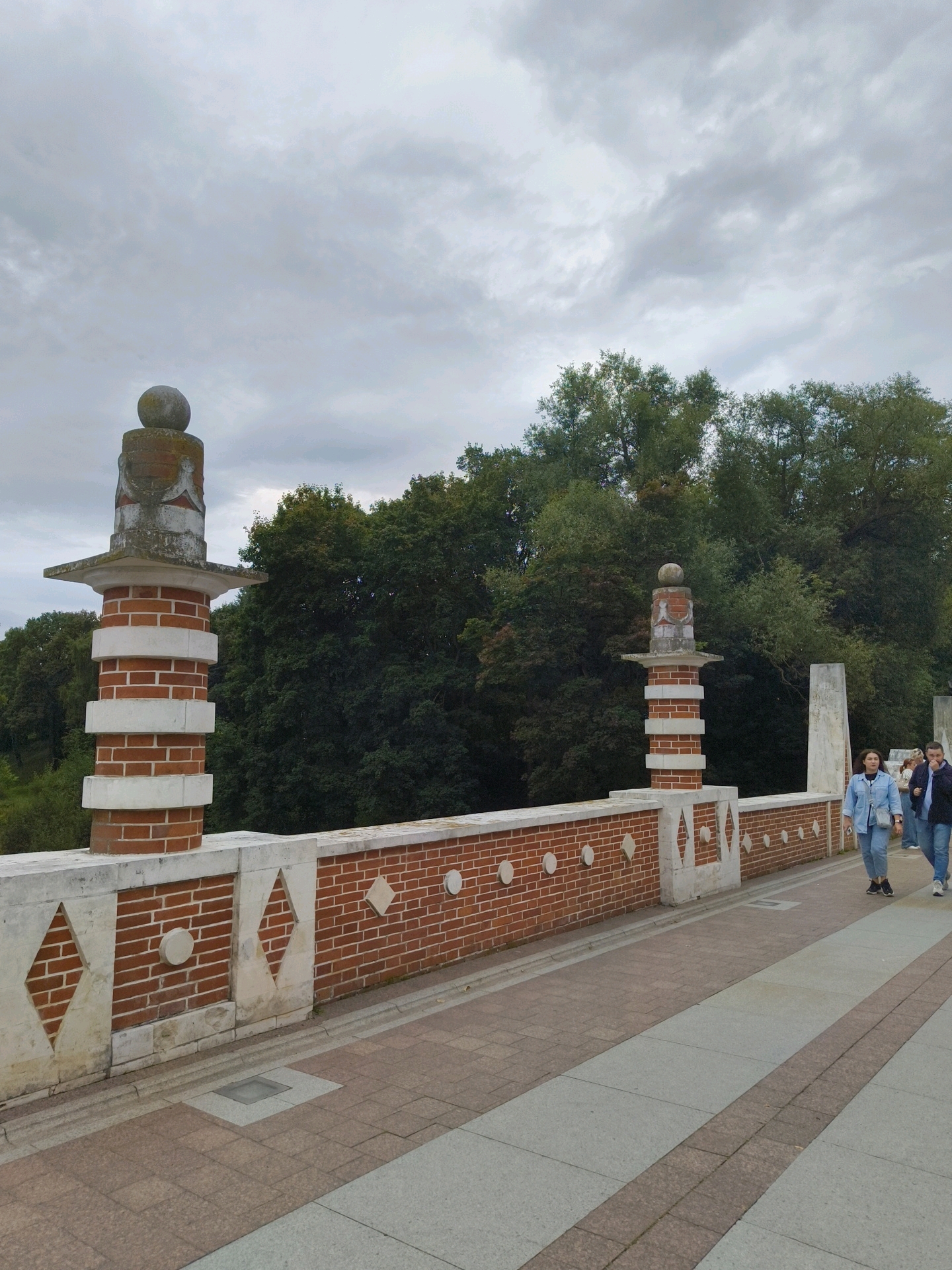 The image shows a brick bridge with decorative pillars in Tsaritsyno Park, Moscow, Russia. The sky is overcast with clouds. The bridge has a red brick railing with white diamond-shaped inlays and circular decorative elements. There are two prominent pillars on the left side of the bridge, made of brick with white bands and topped with stone finials. In the background, lush green trees form a dense forest. On the right side of the bridge, a pathway is visible, with two women and one man walking away from the camera. The woman in the foreground is wearing a denim jacket and jeans. The man next to her is wearing a dark jacket and jeans.