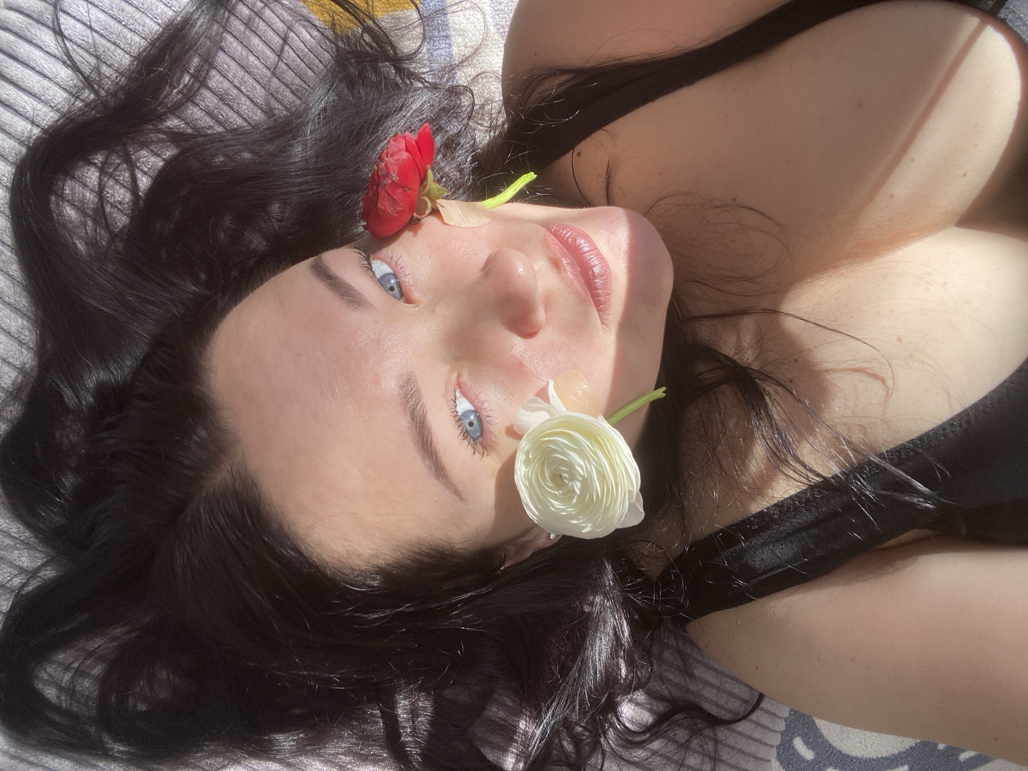 A woman with dark hair lies on a gray and white striped surface. She wears a black top and has a red flower positioned near her forehead and a white flower near her right eye. There is tape on her face. The location of the image is not specified within Sochi, Russia.
