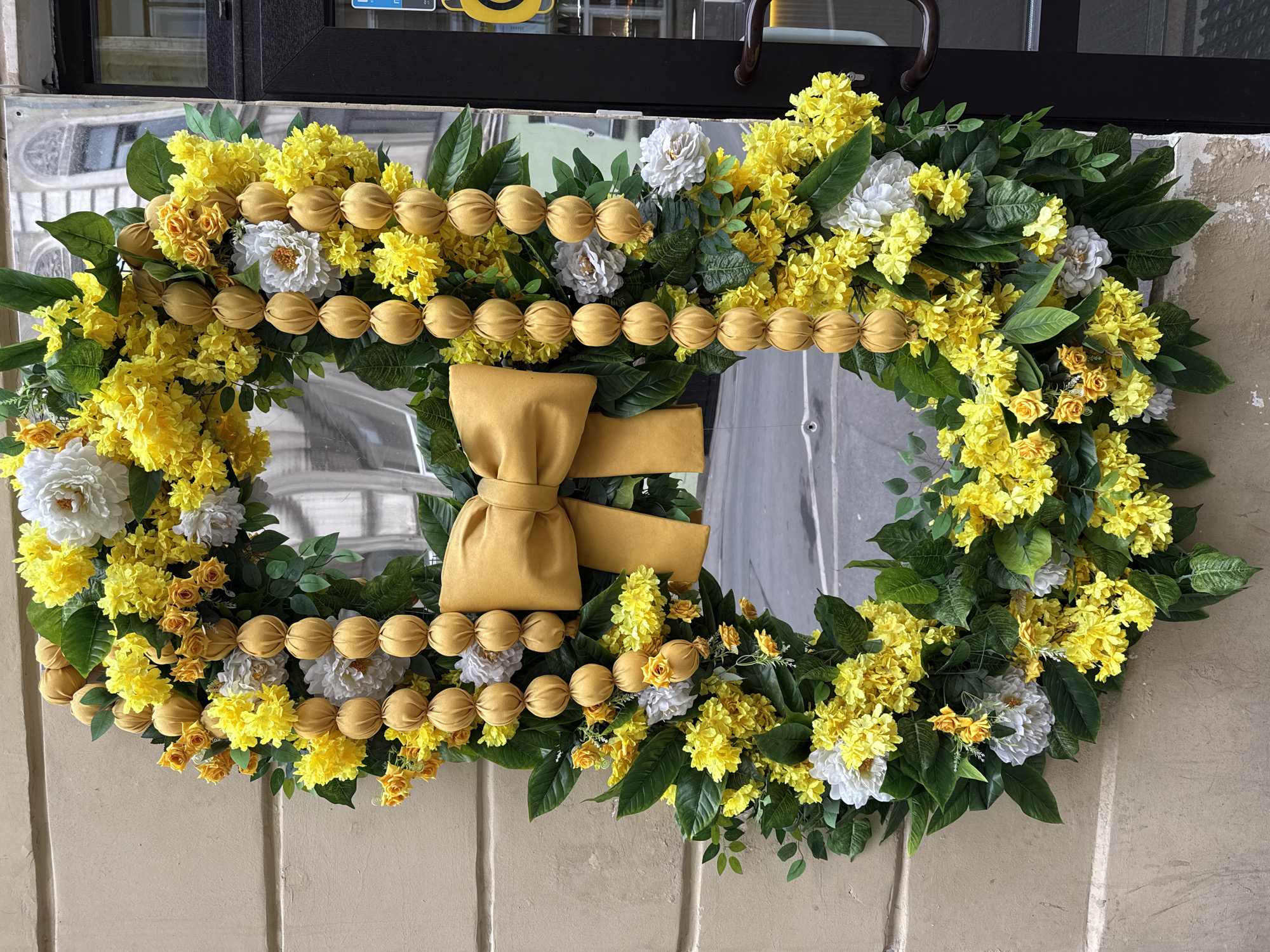 A decorative wreath with yellow and white flowers, green leaves, and a yellow fabric bow is hanging on a mirror outside a building with white siding. The mirror reflects a partial view of a building across the street, suggesting an urban setting. Based on the architectural styles visible in the reflection, this could be in the city of Saint Petersburg, Russia. A dark-colored door frame and handle are visible above the wreath.