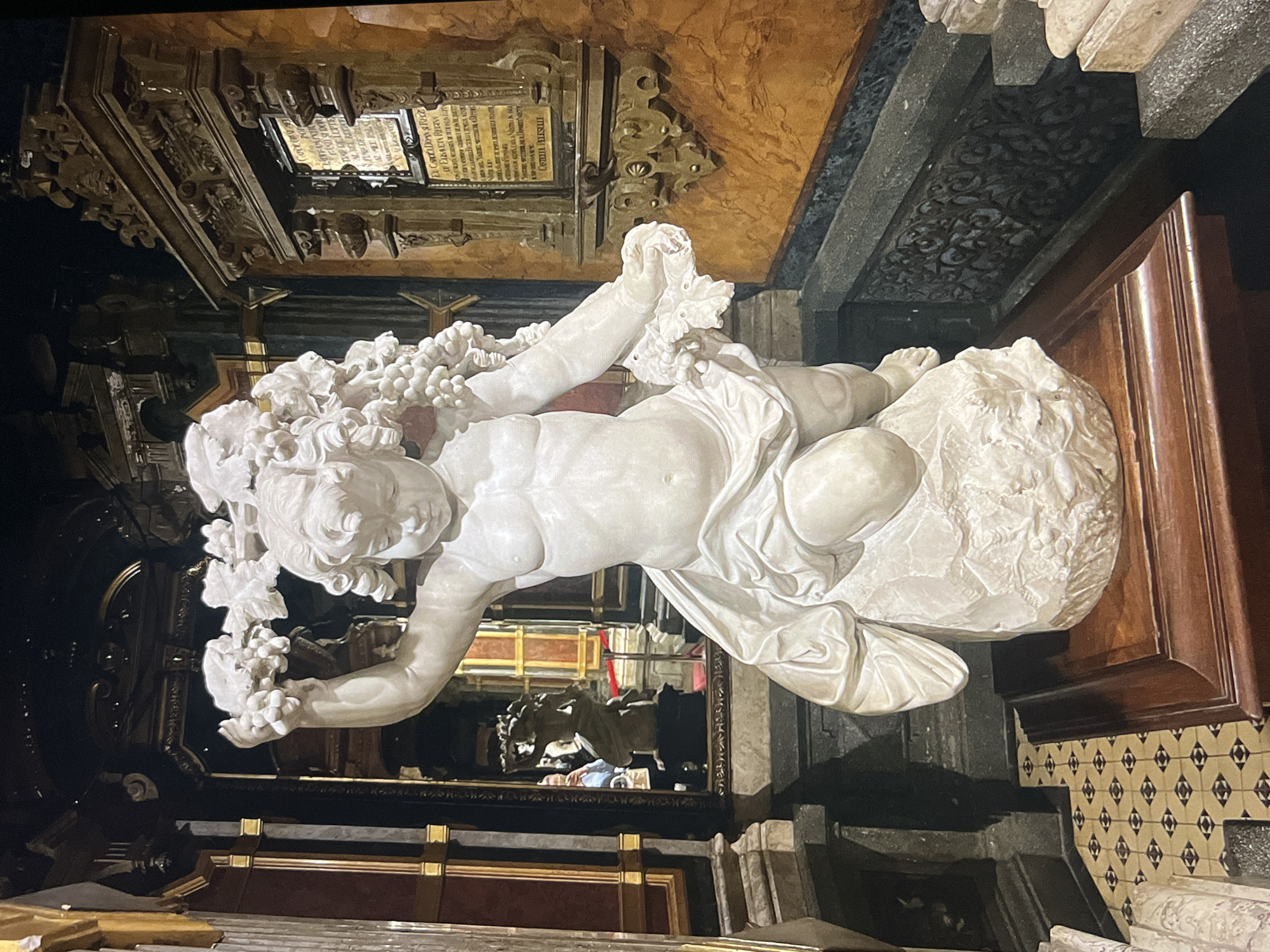 The image depicts a white marble sculpture of a cherubic figure resting on a cloud-like structure, adorned with grapes. The sculpture is part of an elaborate architectural setting, possibly within a church or mausoleum. The background includes ornate wooden structures, marble elements, and decorative tiles. The setting appears to be indoors, well-lit, and of historical or artistic significance. The specific location is Unknown, Unknown.