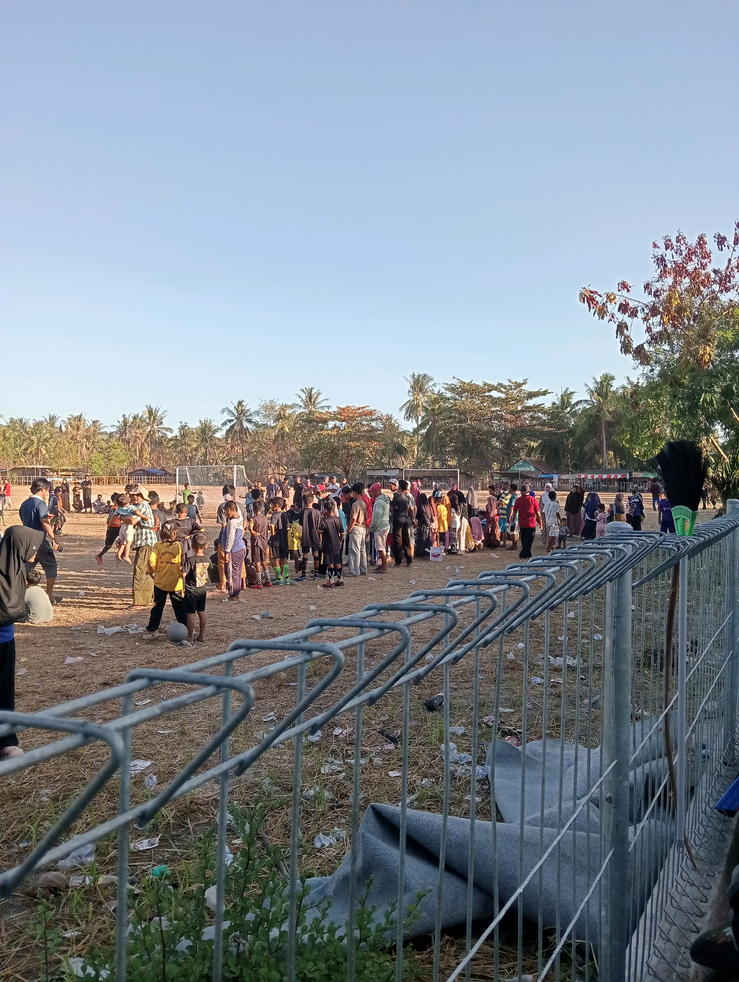 In the city of Pagerungan Kecil, Indonesia, a dusty, dry soccer field is the setting for a gathering of people. The scene is characterized by the presence of a large crowd, predominantly children, some of whom appear to be involved in a soccer-related activity, evidenced by the presence of a soccer ball on the field and a goalpost in the background. The people are dressed in casual attire, with a variety of colors and styles visible. The environment is a sun-drenched outdoor space with palm trees and other vegetation lining the perimeter of the field. A wire fence runs across the foreground of the image, partially obscuring the view of the field and its occupants. Scattered debris, including plastic waste, is visible on the dry, straw-like ground. The sky is a clear, light blue, indicating fair weather. The overall impression is that of a community event or casual sporting activity taking place in a rural or semi-rural Indonesian setting.