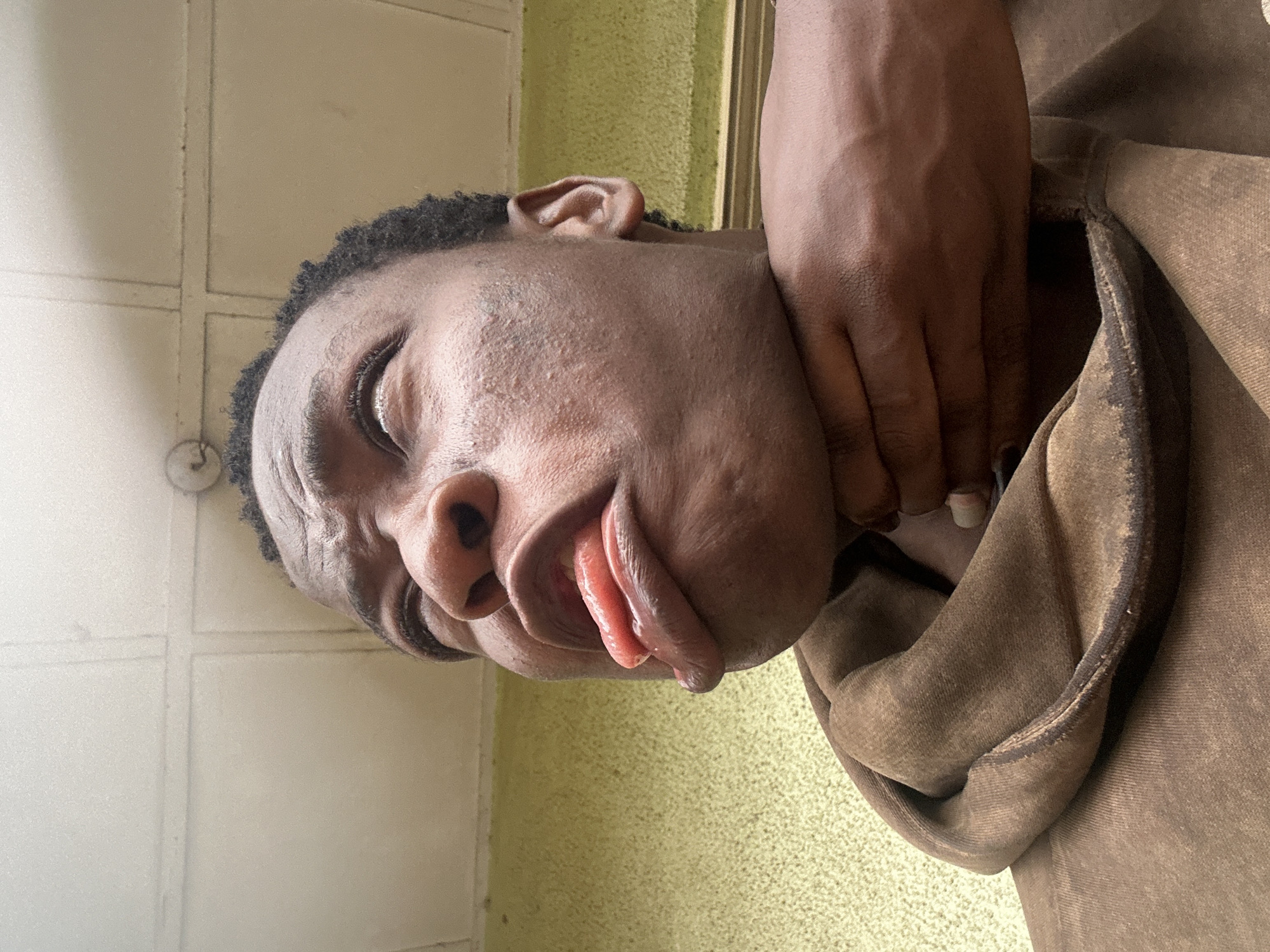 A close-up, rotated portrait shot shows a dark-skinned man with a brown hoodie. His eyes are partially closed, and his tongue is protruding slightly from his mouth. A hand, presumably belonging to another individual, is gripping his neck. The background consists of a tiled wall on the left and a yellow-textured wall on the right. The location is unknown.