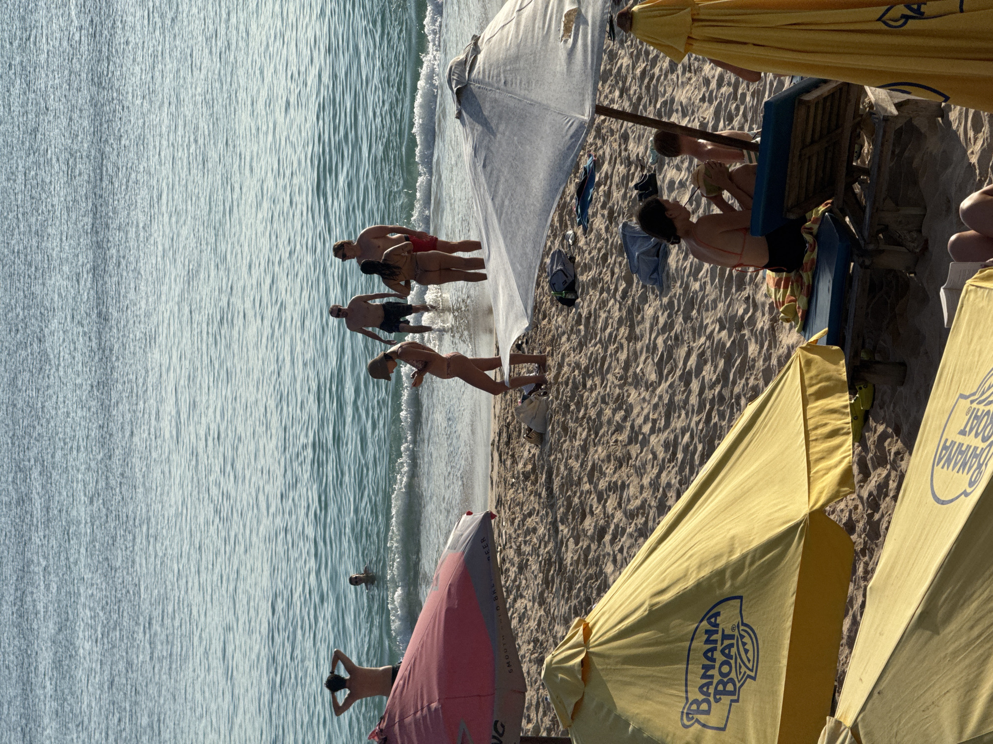 The image captures a beach scene in Kuta Selatan, Indonesia. The ocean is on the left side with gentle waves. Several people are wading in the water. There are four adults and one child. A beach with golden sand occupies the right side. Two yellow beach umbrellas, one prominently displaying the "Banana Boat" logo, dominate the lower right foreground. In the upper part of the image are other people on lounge chairs.