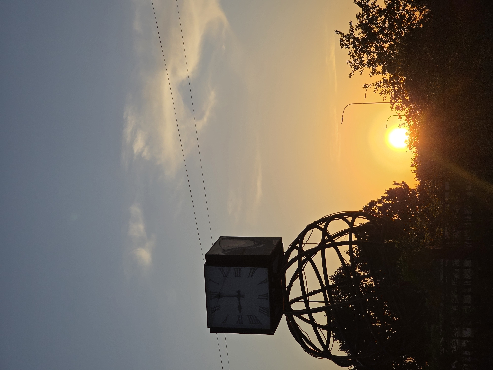 The image depicts a clock tower, silhouetted against a sunset sky in Chaikovsky, Russia. The clock is square with Roman numerals. It is attached to a spherical structure made of interconnected metal bands. Power lines are visible against the sky. The sun is obscured by trees and the branches are dark silhouettes. A lamppost is also silhouetted next to the sun. The sky is colored blue at the left of the image transitioning to yellow at the right.