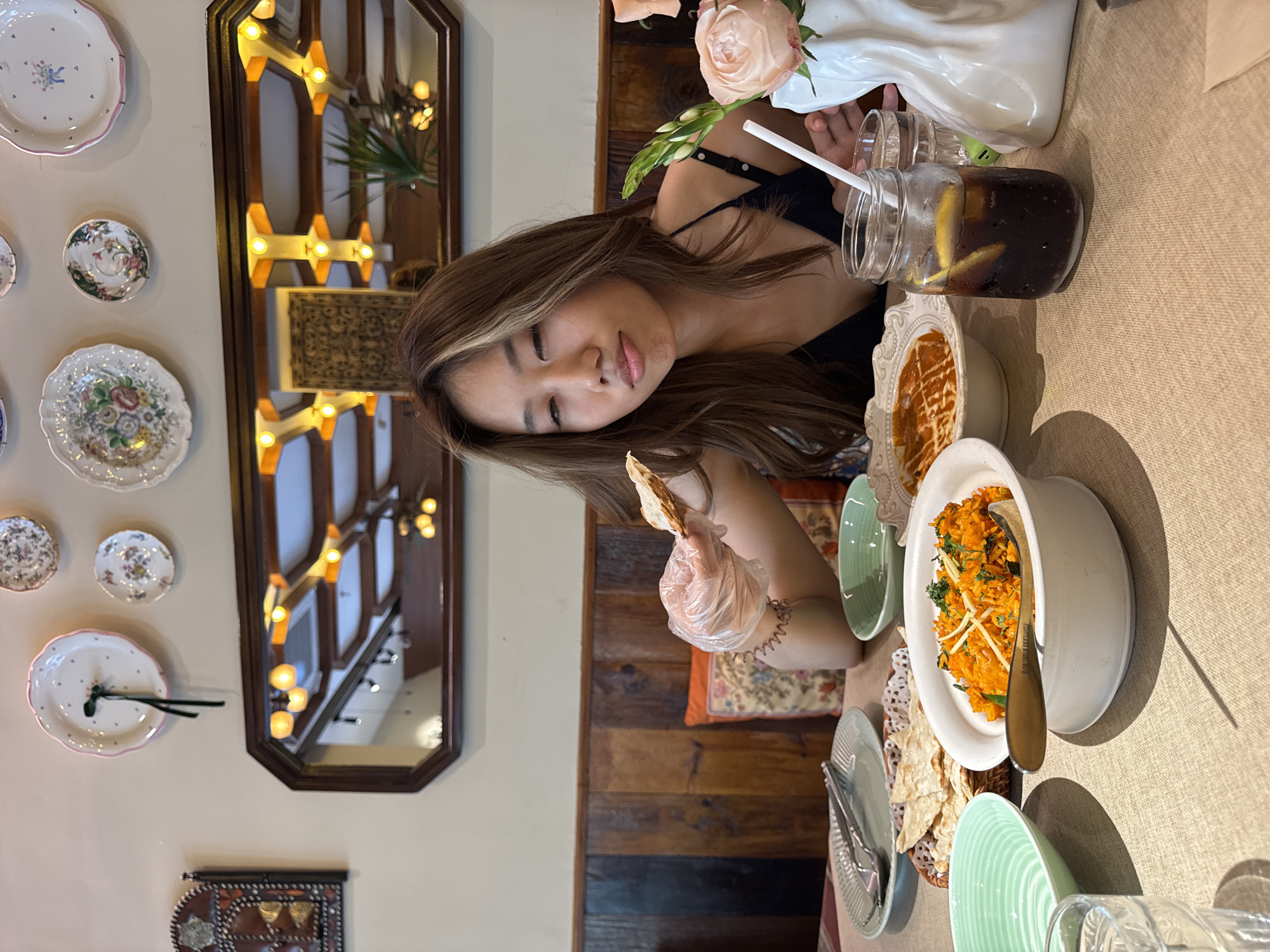 Here is a description of the image:
An overhead shot shows a young Asian woman seated at a table in what appears to be a restaurant. She wears a black top and has long, light brown hair with blonde highlights. She is smiling faintly with her eyes closed and holds a piece of food in her gloved hand. A selection of Indian dishes sits on the table, including rice, dips, and bread. A glass jar containing a dark liquid with lemon slices and a straw, along with a vase of peach-colored roses, is also on the table.
The setting is an interior space with a unique aesthetic. A mirror with decorative lights reflects the restaurant's interior. Decorative plates hang on the wall, and the seats have patterned cushions. Wood paneling lines part of the wall. While the image does not give specific landmarks, there are indicators it could be a stylized, themed restaurant, but it does not provide explicit details that would confirm a location in Yuzhong District, China.