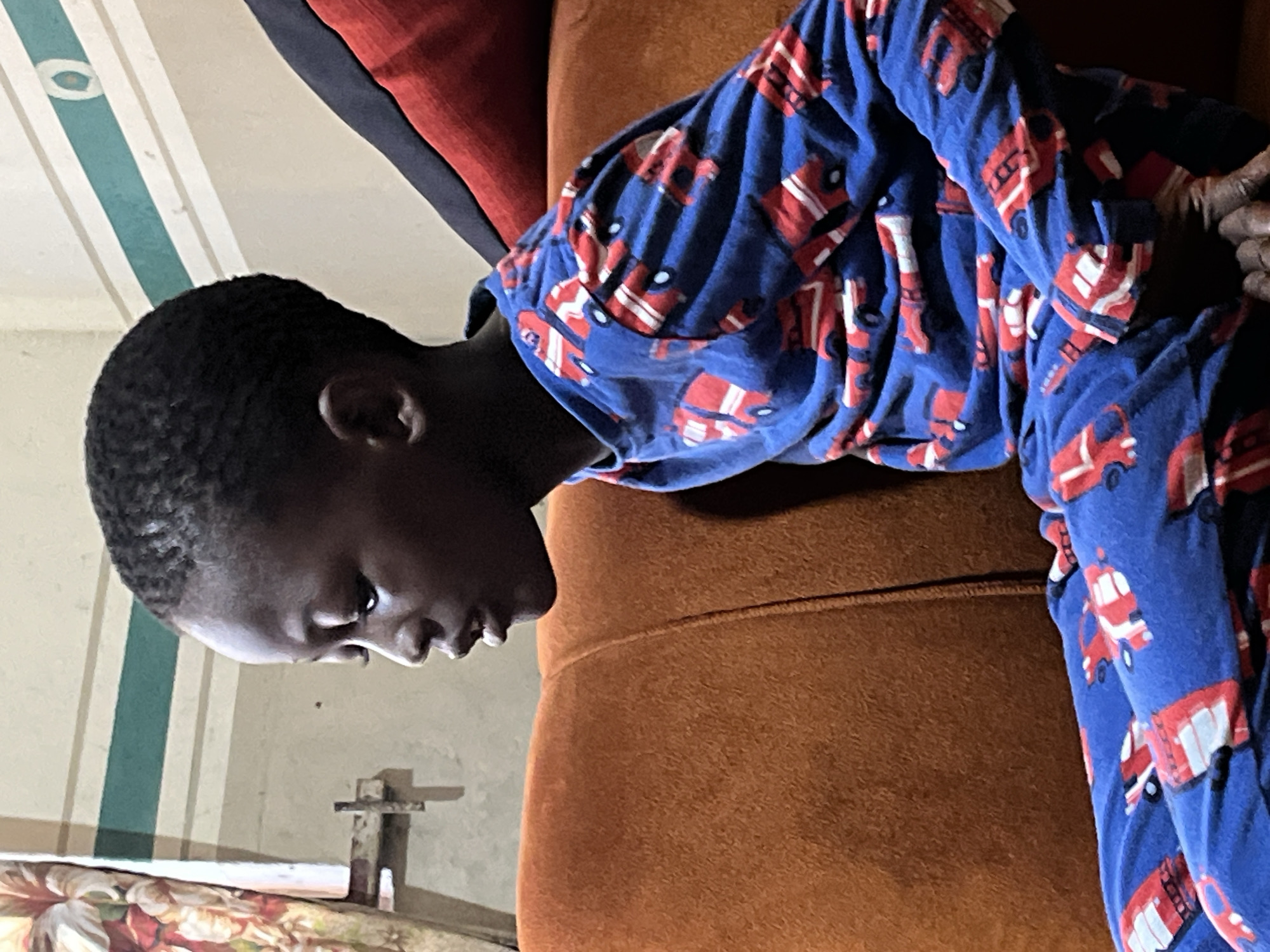 A child in blue pajamas with a fire engine pattern is lying face down on an orange couch. The background includes a wall with a green and white striped pattern and a floral design visible near the bottom. The location is not discernible as Badagry, Nigeria based on the information in the image.