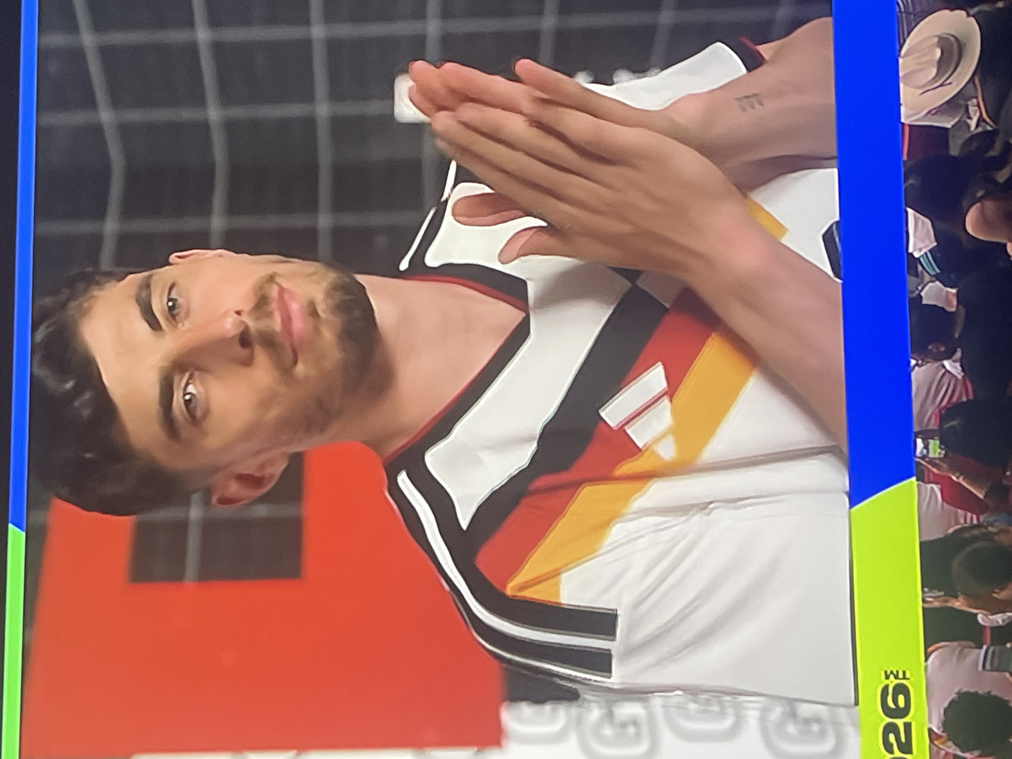 The image shows a man wearing a white, black, and red German national football team jersey. He is clapping his hands together in front of him. The man has short, dark hair, a light beard, and is looking slightly off-camera. In the background, there is a red wall on the left and a black grid pattern on the right. There is a green and blue banner at the bottom of the image, with some text visible. The scene is indoors, possibly a studio setting or a media event, and there are blurred figures of people in the lower part of the image, suggesting an audience or spectators. There is no discernible indication of the city of Ruse, Bulgaria, within the image itself.