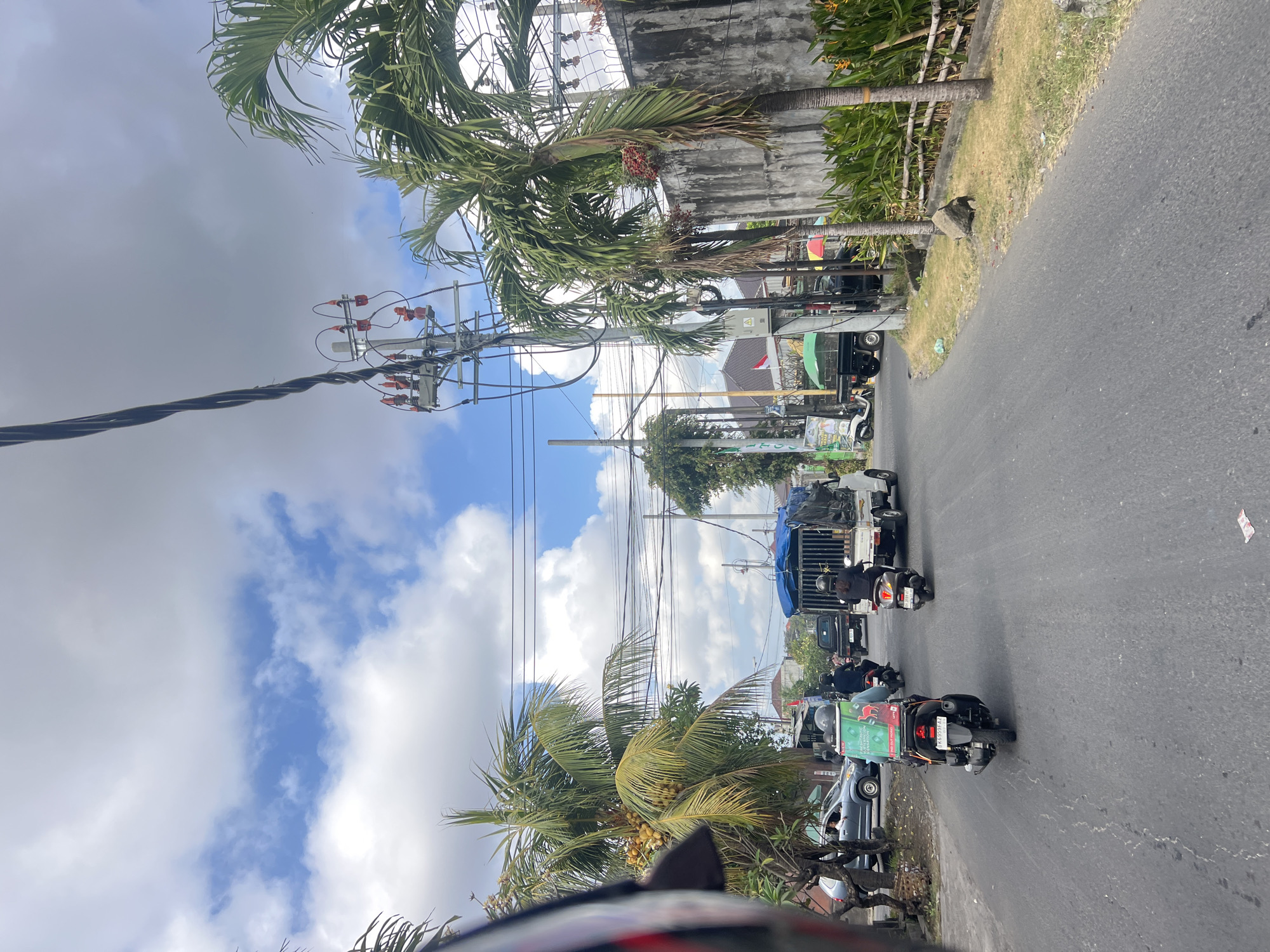 This is a medium-shot, outdoor, street-level, eye-level perspective photograph taken on a paved road in Denpasar, Indonesia. The scene depicts moderate traffic under a partly cloudy sky.
In the foreground, a person on a black scooter, with a green and red box on the back, is visible. To the right of this scooter, another person on a black scooter is also heading forward. A white truck with a blue tarp covering its load is in front of them, also moving forward. Further ahead on the left, a grey car is visible, with at least one person inside. In the background, various buildings, palm trees, and electrical poles with wires are present. The road surface shows signs of wear. The overall environment suggests a typical urban street scene in a tropical climate.