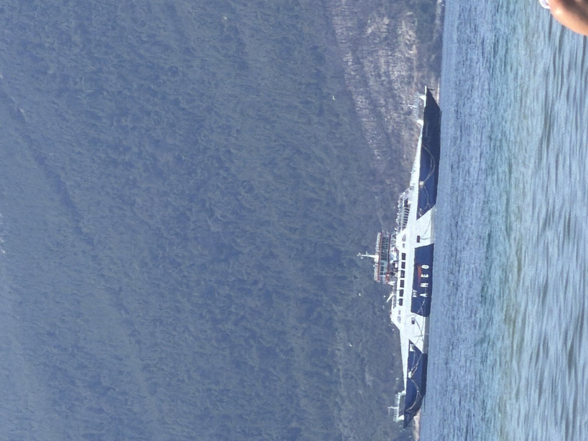 A wide shot captures a large white ferry with blue accents navigating a calm body of water. The ferry displays the text "ANEO" in large blue letters on its side. In the background, a densely forested, green mountainside rises steeply. The sky above is a light blue. The foreground features the rippling surface of the water, with a hint of a beach or shoreline on the right edge, where a person's arm is partially visible. The scene is set near the city of Thassos, Greece.