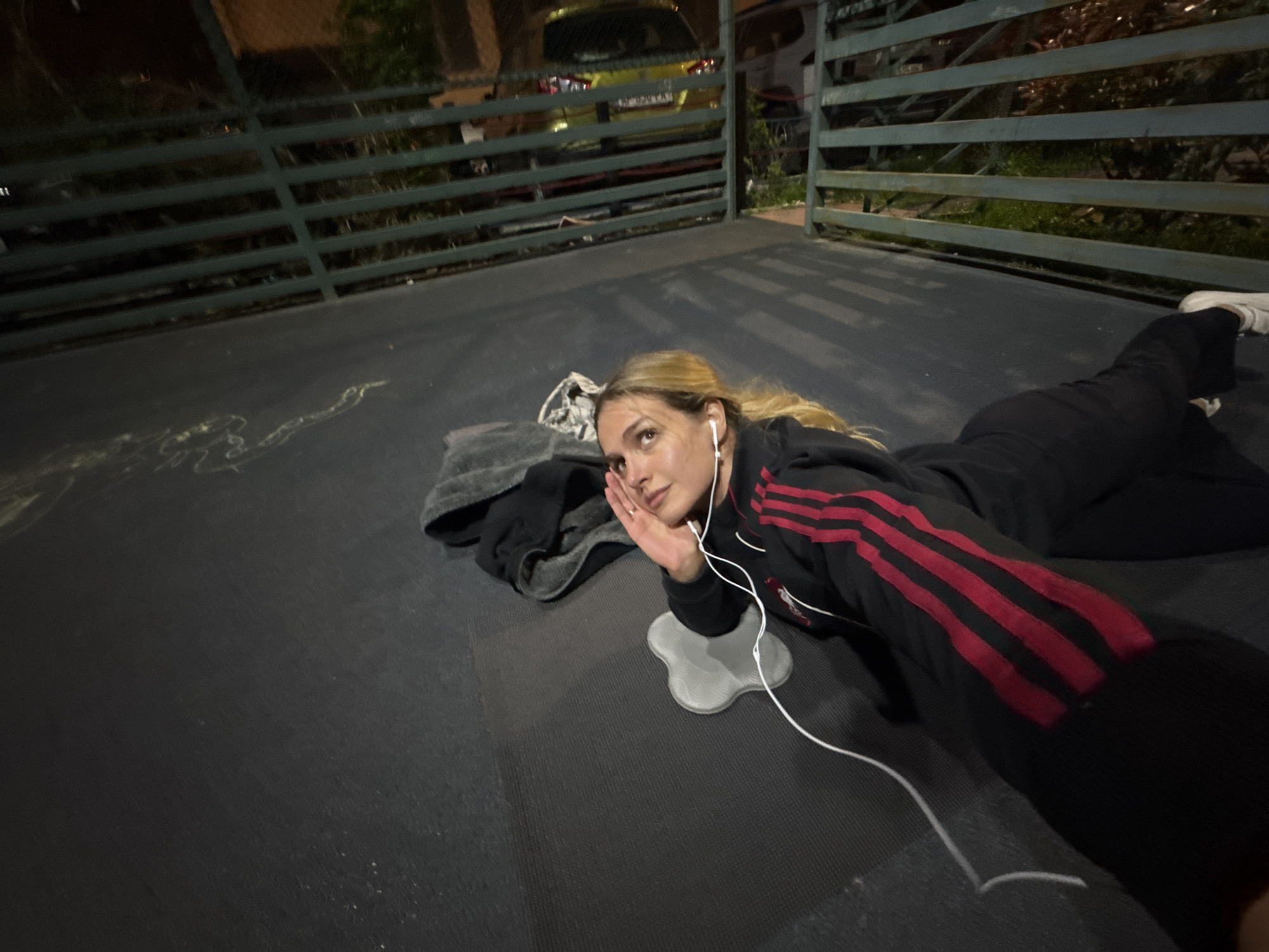 A woman lies on a dark floor in what appears to be an outdoor public space in Tbilisi, Georgia, at night. She wears a black tracksuit with red stripes down the arm, and white earbuds are connected to a device out of frame. She is resting her head on her hand, and a small, gray, flower-shaped pad is beneath her head. A dark gray blanket is to her left. In the background, a green metal railing runs across the scene. A yellow car is parked beyond the railing. Chalk drawings can be seen on the floor.