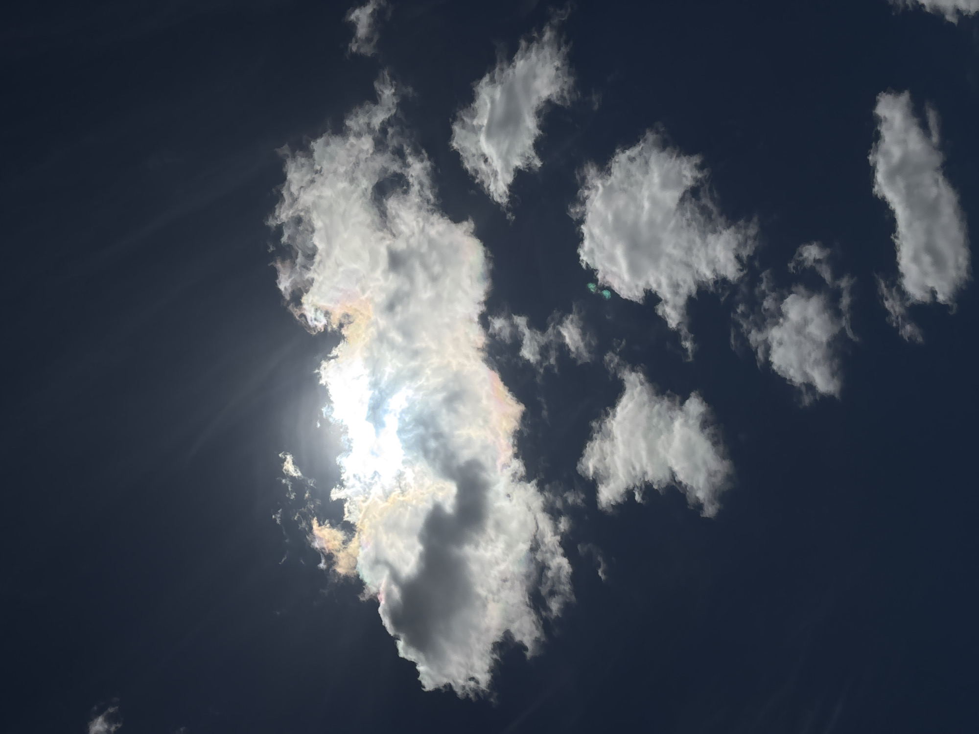 This upward-facing photograph captures a dynamic sky scene over Красные Баки, Russia, dominated by deep blue hues. A large, bright, white cloud formation occupies the left-center of the frame, with the sun's intense light bursting from behind it, creating prominent sunrays. The edges of this main cloud display striking iridescence, with vibrant rainbow-like colors including yellow, orange, pink, and green, a phenomenon caused by light diffraction through tiny ice crystals or water droplets.
Scattered across the dark blue expanse are several smaller, white, cumulus-like clouds, varying in shape and brightness. A couple of distinct green lens flares are visible in the sky, particularly near the main cloud and further to the right, indicating the sun was directly or indirectly in the camera's view. The image depicts a clear daytime sky with scattered clouds, showcasing natural atmospheric optics. There are no people, man-made objects, or visible text in the frame.