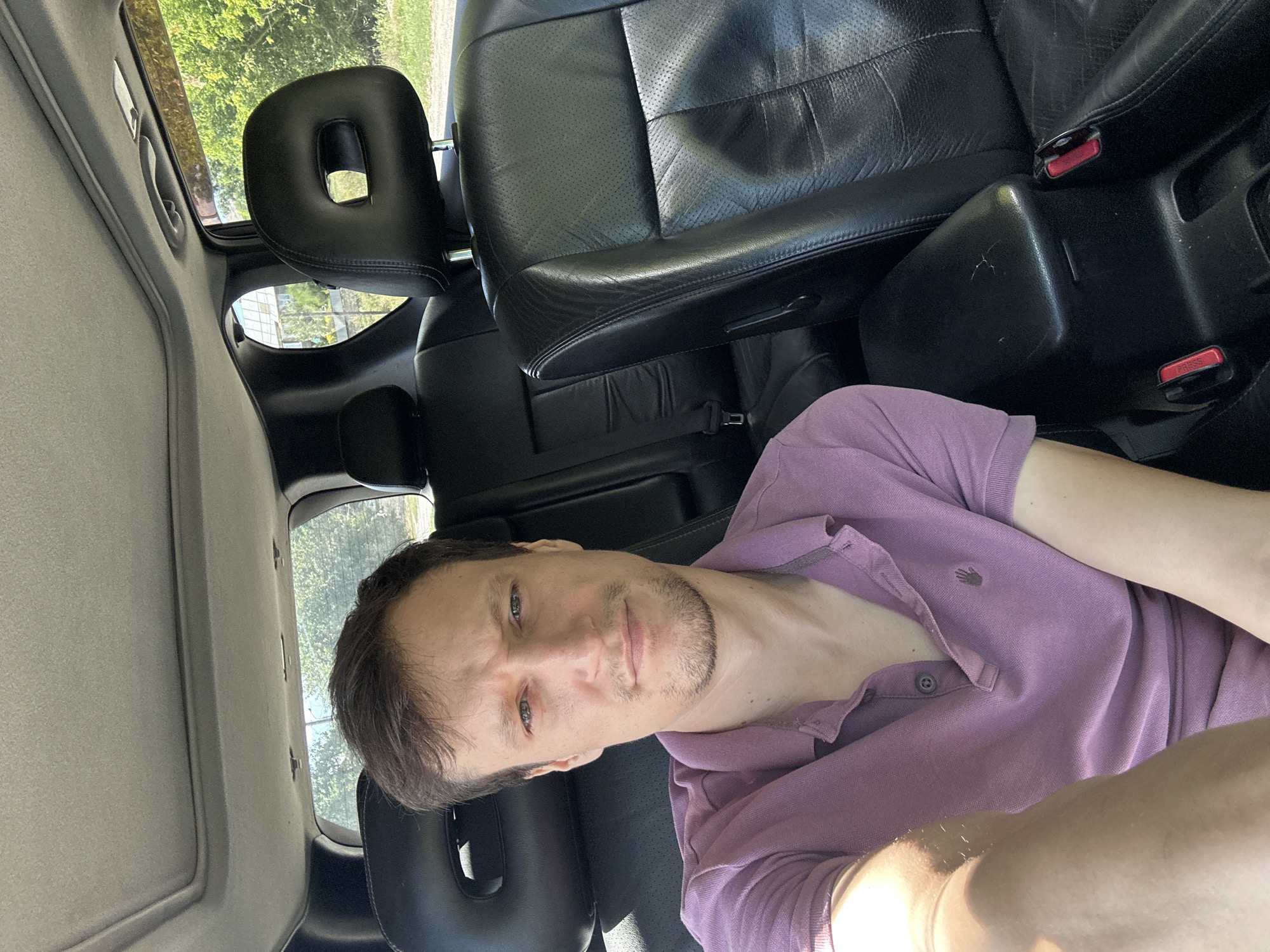 A man with short brown hair and light skin, wearing a pink polo shirt, sits in the back seat of a car. He is looking directly at the camera. The interior of the car is visible, with black leather seats and a sunroof. Outside the car, trees and greenery can be seen. The scene appears to be in a rural or suburban area, possibly on a sunny day.