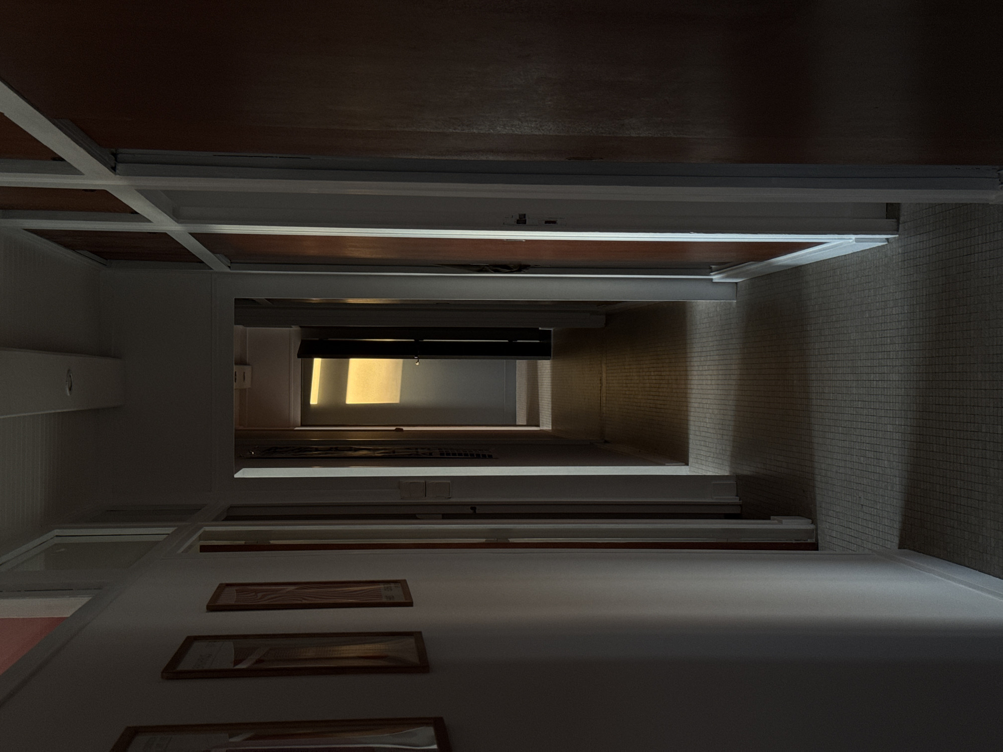 A low-angle view of a hallway looking upward through several floors. The walls are primarily white with some sections of brown paneling and tiled surfaces. A sequence of framed pictures adorns the wall on the left side of the frame. The light source emanates from a window or opening at the end of the hallway. The location is Soulac-sur-Mer, France.