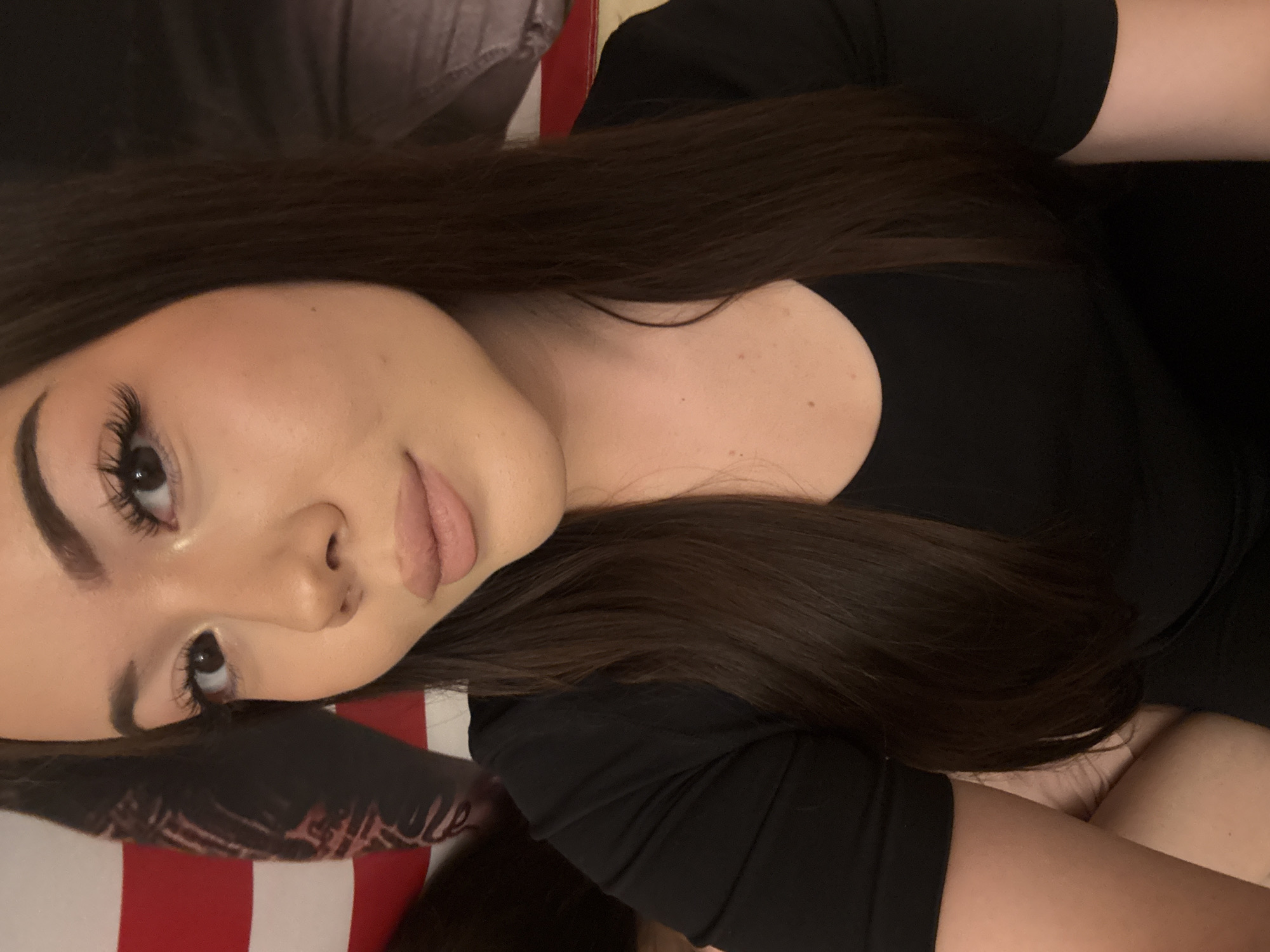 This media file is an indoor portrait of a young woman, potentially taken in Naberezhnye Chelny, Russia. She is depicted lying on her side, with her head resting on a surface, looking subtly towards the viewer. Her long, straight, dark brown hair flows around her. She is wearing a black garment, likely a top or dress. Her complexion is fair, with several small moles visible on her shoulder and upper chest area.
Her facial features are prominent: she has long, dark eyelashes, likely enhanced by makeup or extensions, and meticulously shaped, dark eyebrows. Her eyes are dark brown, and her lips are a light, natural shade. She has a serene and composed expression, with a hint of a smile.
In the background, behind her head and extending beneath her, is a textile with a distinct red and white striped pattern, suggesting bedding or a cushion. Part of a lighter, possibly cream-colored, blanket or pillow is also visible. Her right arm is bent and partly visible, as is a portion of her upper leg, suggesting she is reclining comfortably. The lighting appears soft and uniform, typical of an indoor setting. There is no visible text in the image.