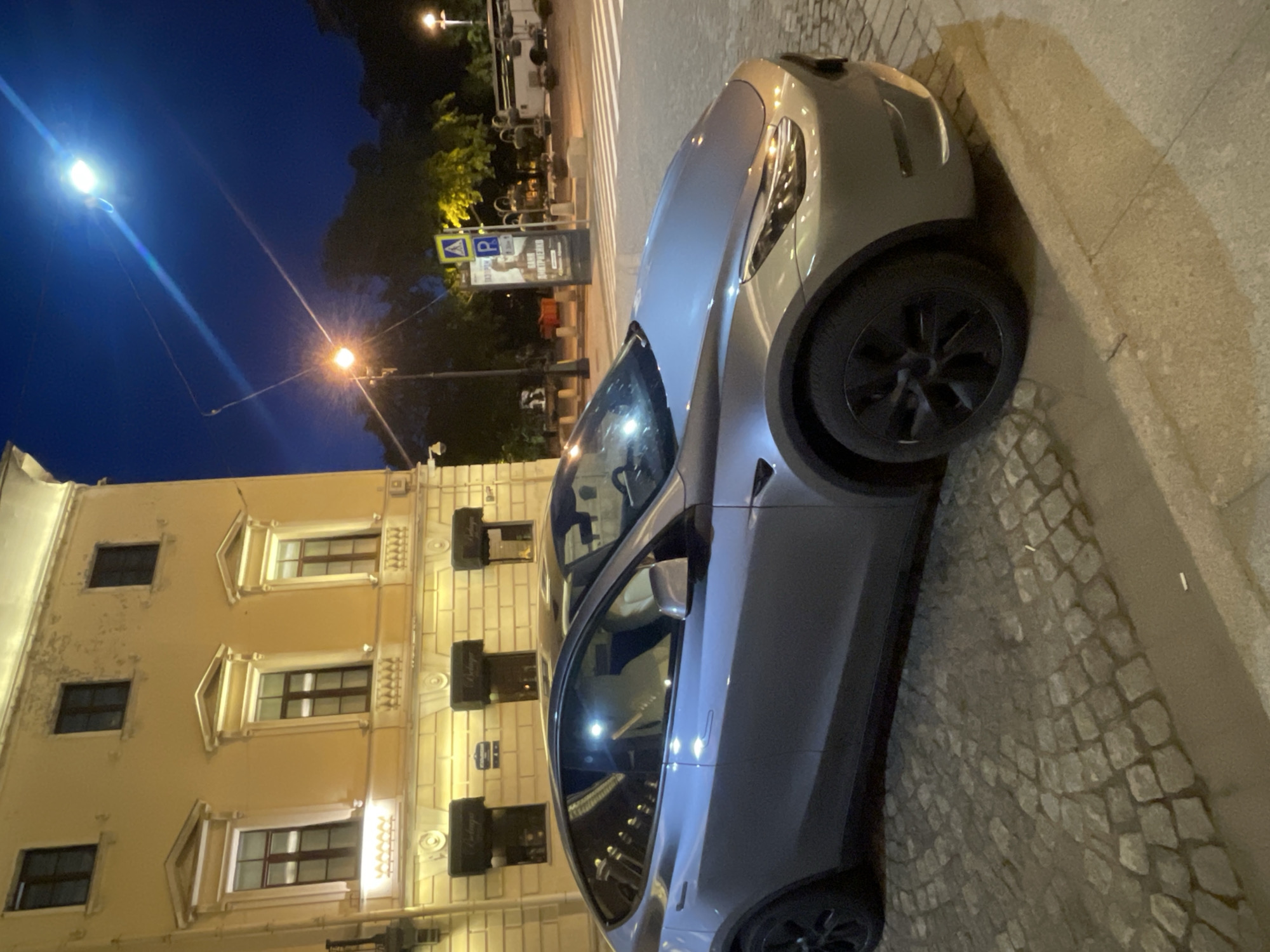 This image captures a night scene on a cobblestone street in Saint Petersburg, Russia. The primary subject is a silver-gray Tesla, likely a Model Y, parked on the right side of the frame, angled towards the upper left. Its distinctive black wheels and sleek body are prominent under the streetlights.
Adjacent to the car on the left is a multi-story, light-colored building with classic architectural details, including decorative window frames and cornices. Several windows are visible, some illuminated from within. Attached to the building are small black signs, one of which clearly reads "Bodega" and "winebar & tapas," indicating a business on the ground floor.
The sky is dark blue, typical of nighttime. Bright streetlights punctuate the scene; a strong, cool-toned light source is prominent on the upper left, while a warmer, yellow-orange light is visible in the middle-ground. In the background, behind the car, a blue square sign with a white "P" and an arrow (indicating parking) is visible, alongside a yellow triangular sign with a black pedestrian symbol (a crosswalk warning). A billboard or poster featuring a person is partially visible behind these signs.
The street is paved with distinctive cobblestones, and a concrete curb or sidewalk runs along the right edge of the frame. No people are visible, and no specific activity is taking place beyond the car being parked. The overall ambiance is quiet and illuminated by artificial light, showcasing the urban architecture and a modern vehicle in a historical setting. The image is rotated 90 degrees clockwise.