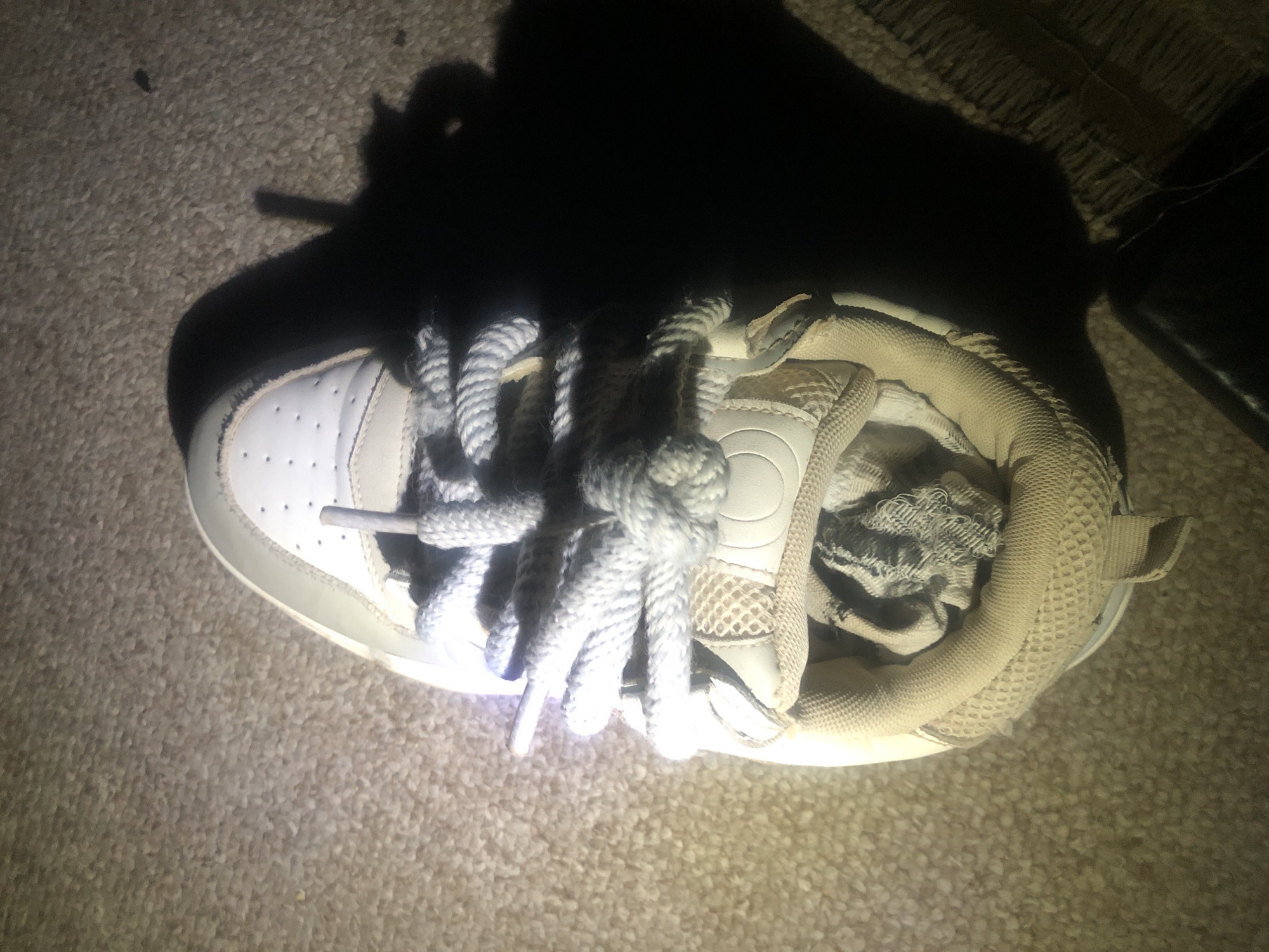 An overhead view of a single, light-colored sneaker is captured on a textured, beige carpet. The sneaker is positioned vertically in the frame, with the toe pointing upwards. Thick, light-grey laces are prominently displayed, tied in a knot and looping through eyelets. The shoe's interior is partially visible, revealing a crumpled, greyish fabric. A dark shadow is cast to the right of the sneaker, obscuring a portion of the carpet. The lighting appears to be artificial and directional, creating strong contrasts. This scene does not provide any specific information about the city of Jalingo, Nigeria, or any people or actions taking place.