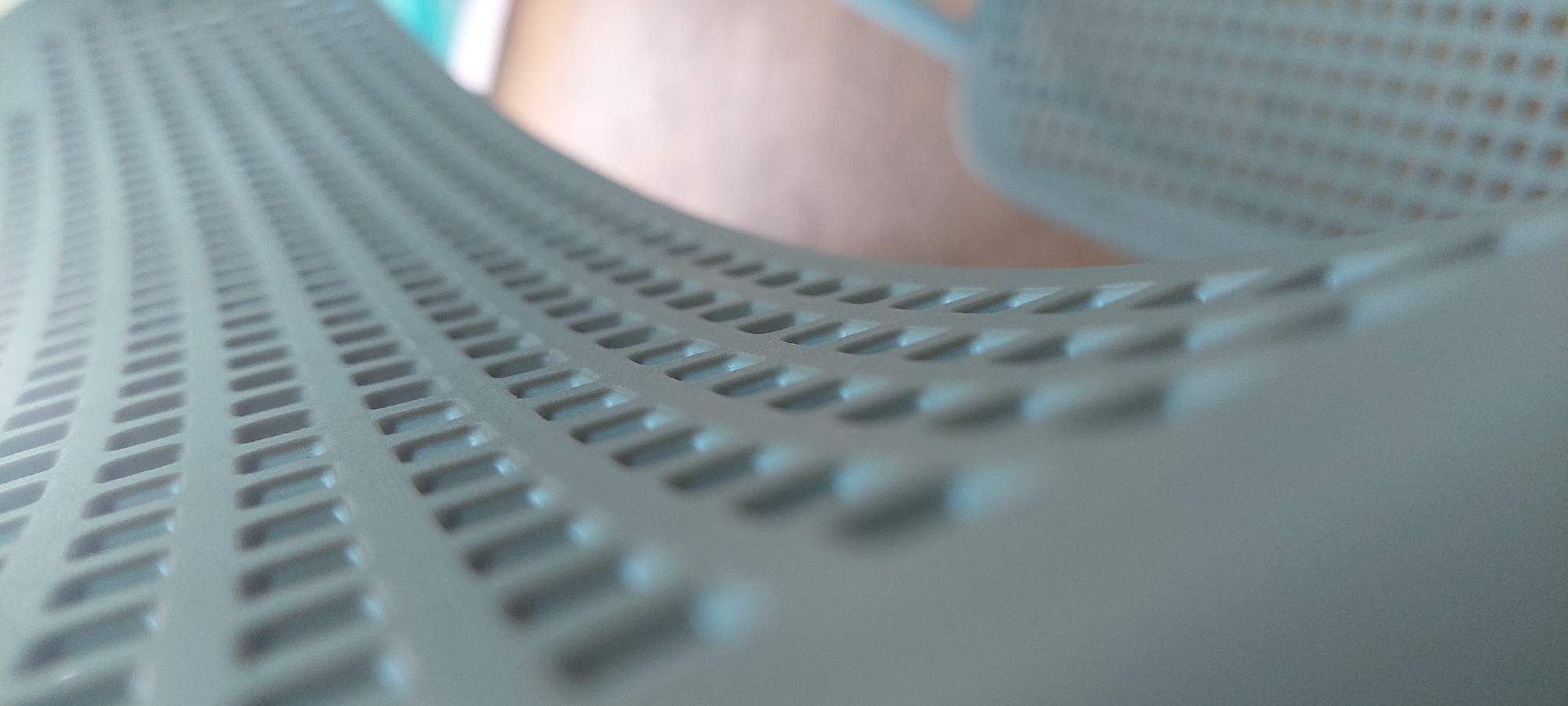 The image is a close-up, abstract shot of a perforated, light-colored chair, likely made of plastic. The camera angle is low and angled, focusing on the curved surface of the chair and its repeating pattern of rectangular perforations. The perforations create a sense of depth and texture, with light and shadow playing across the surface. The background is blurred and out of focus, suggesting an indoor environment with a wooden floor and possibly a light-colored wall or another surface. There are no people or identifiable landmarks visible. The image was taken in Berod bei Hachenburg, Germany.