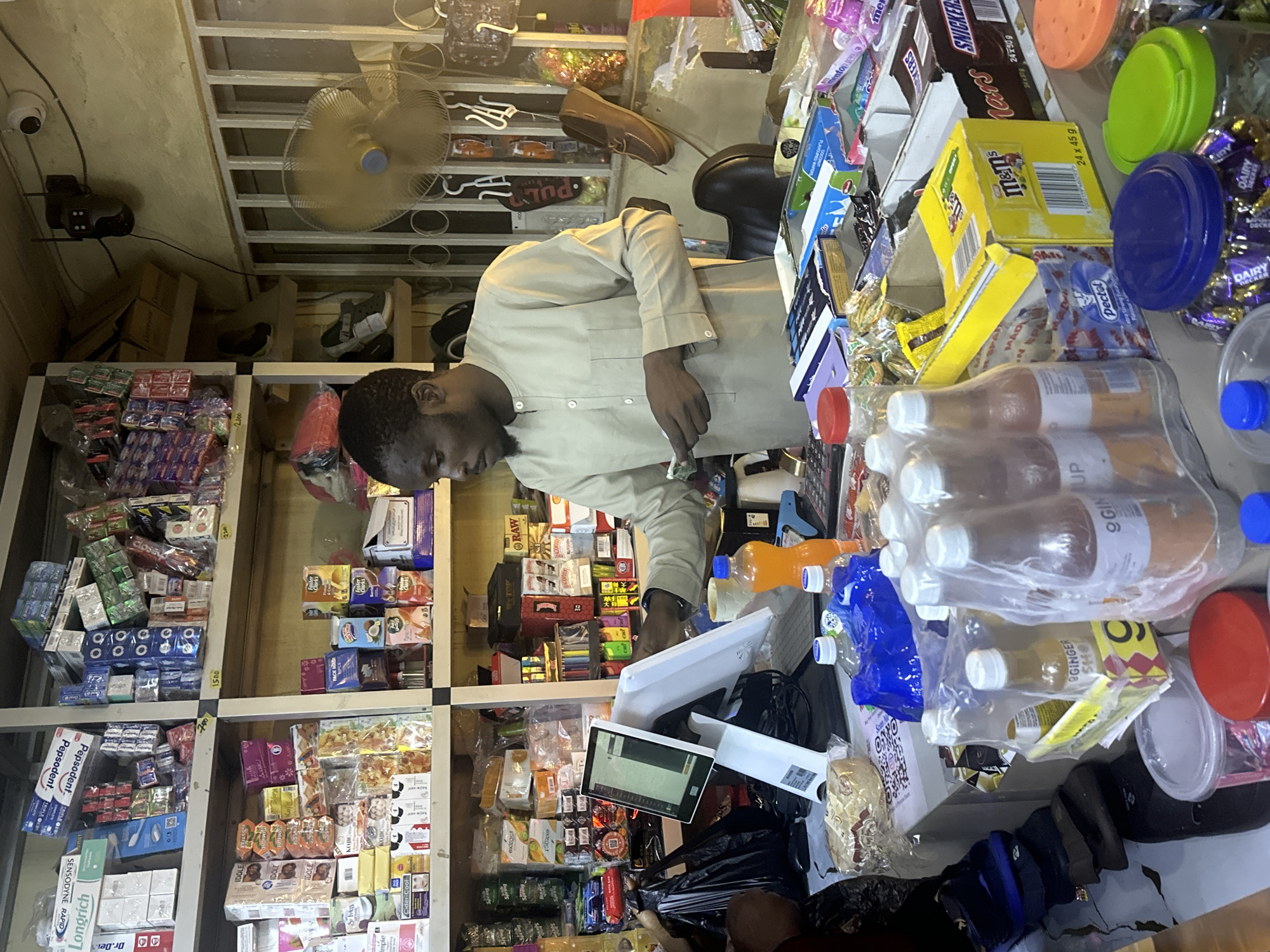This is a full, eye-level shot of a small convenience store in Abuja, Nigeria, with a male shopkeeper interacting with a customer, who is mostly out of frame. The shopkeeper, a dark-skinned man wearing a light beige kaftan, stands behind a counter displaying various goods. He is holding a few Nigerian Naira notes and appears to be making a transaction.
The counter is laden with products: several bottles of what appear to be ginger drinks and other beverages, a large blue plastic bag filled with water bottles, and a white packet of biscuits. To the right of the shopkeeper, a white electronic point-of-sale terminal is visible. The shelves behind the counter are packed with a diverse array of products, including toiletries, packaged snacks, and drinks.
On the wall behind the counter, a white ceiling fan is mounted, and a black security camera is visible in the upper right corner. The store appears to be dimly lit, typical of many shops in the region. The shelves are constructed from white metal and wood. The overall impression is that of a busy, well-stocked local shop catering to everyday needs.