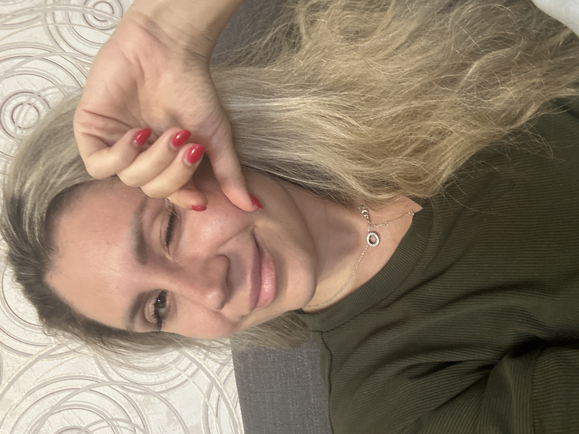 A medium shot shows a woman with blonde hair and red fingernails winking. She is wearing a dark green, ribbed shirt and a silver necklace with a circular pendant. The background consists of a grey couch and textured wallpaper with a circular pattern. The lighting is soft. No specific location within Novoaltaysk, Russia, can be determined from the image.