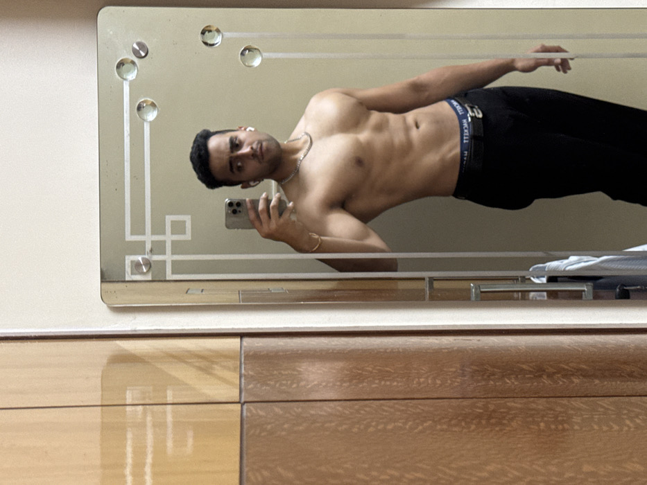 A shirtless man with dark hair, dark pants, and a silver necklace is shown reflected in a vintage mirror. He is holding a phone to take a selfie. The mirror has decorative glass circles and a geometric pattern. Below the mirror is a wooden dresser with varied tones.