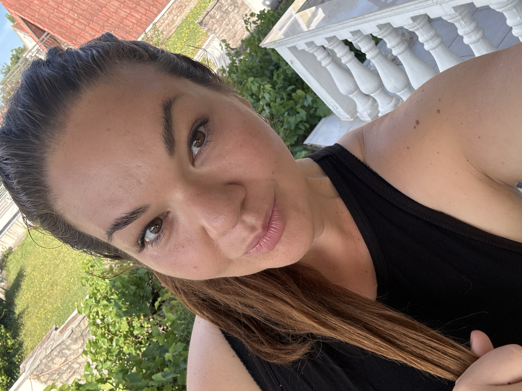 A selfie depicts a woman with fair skin, brown hair, and brown eyes, posing outdoors. She wears a black tank top. Behind her, there is green foliage and a white decorative railing, suggesting a balcony or porch. In the distance, a red-tiled roof can be seen. Based on the prompt, the location is Bijela, Montenegro.